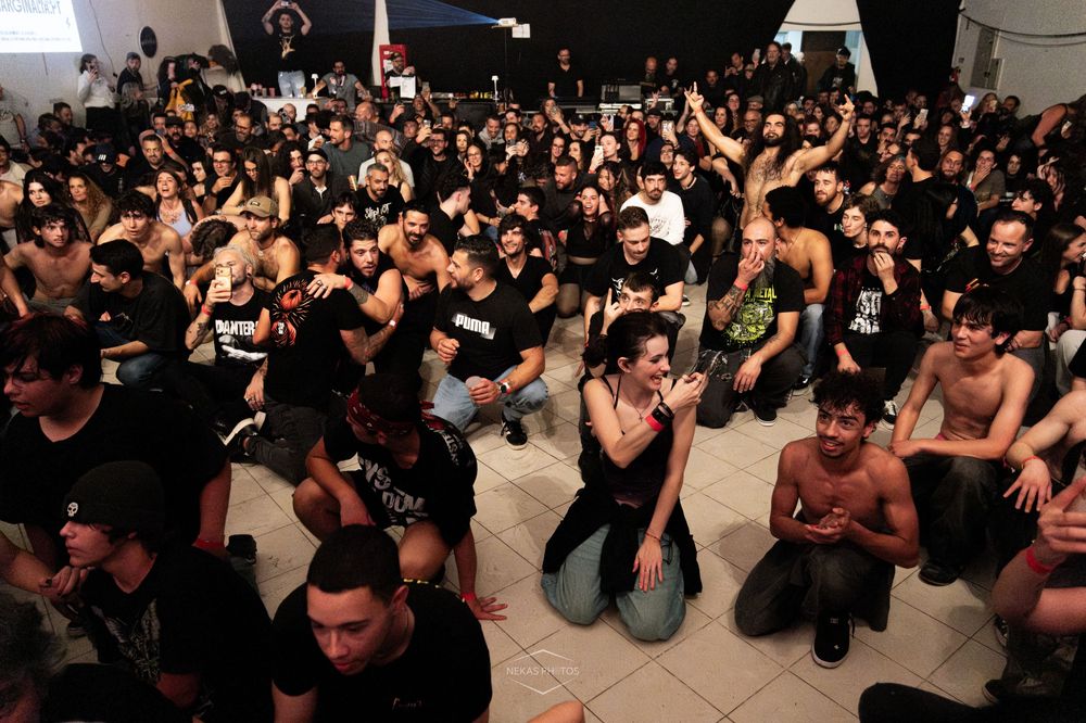 Large crowd of people in black clothing sitting and standing at an indoor concert or music event venue.