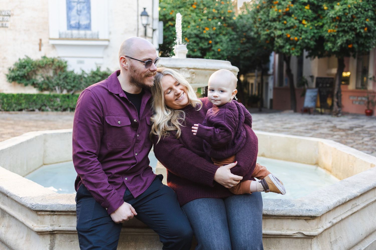A happy family of three in burgundy outfits sits by a stone fountain, parents smiling at their baby outdoors.