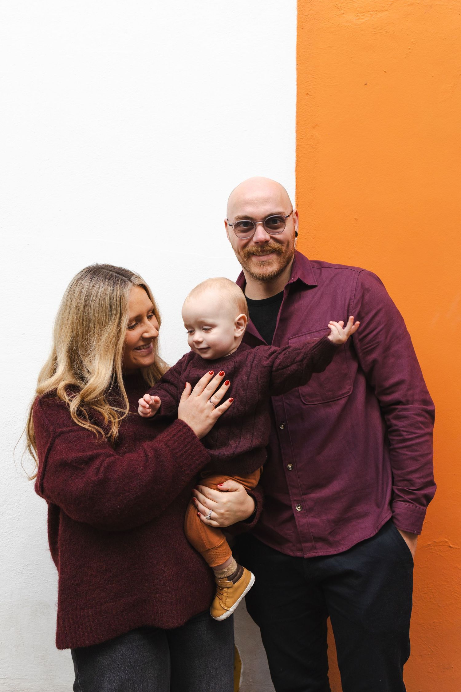 A young family with a bald father, blonde mother, and baby in burgundy outfits pose against a white and orange wall.