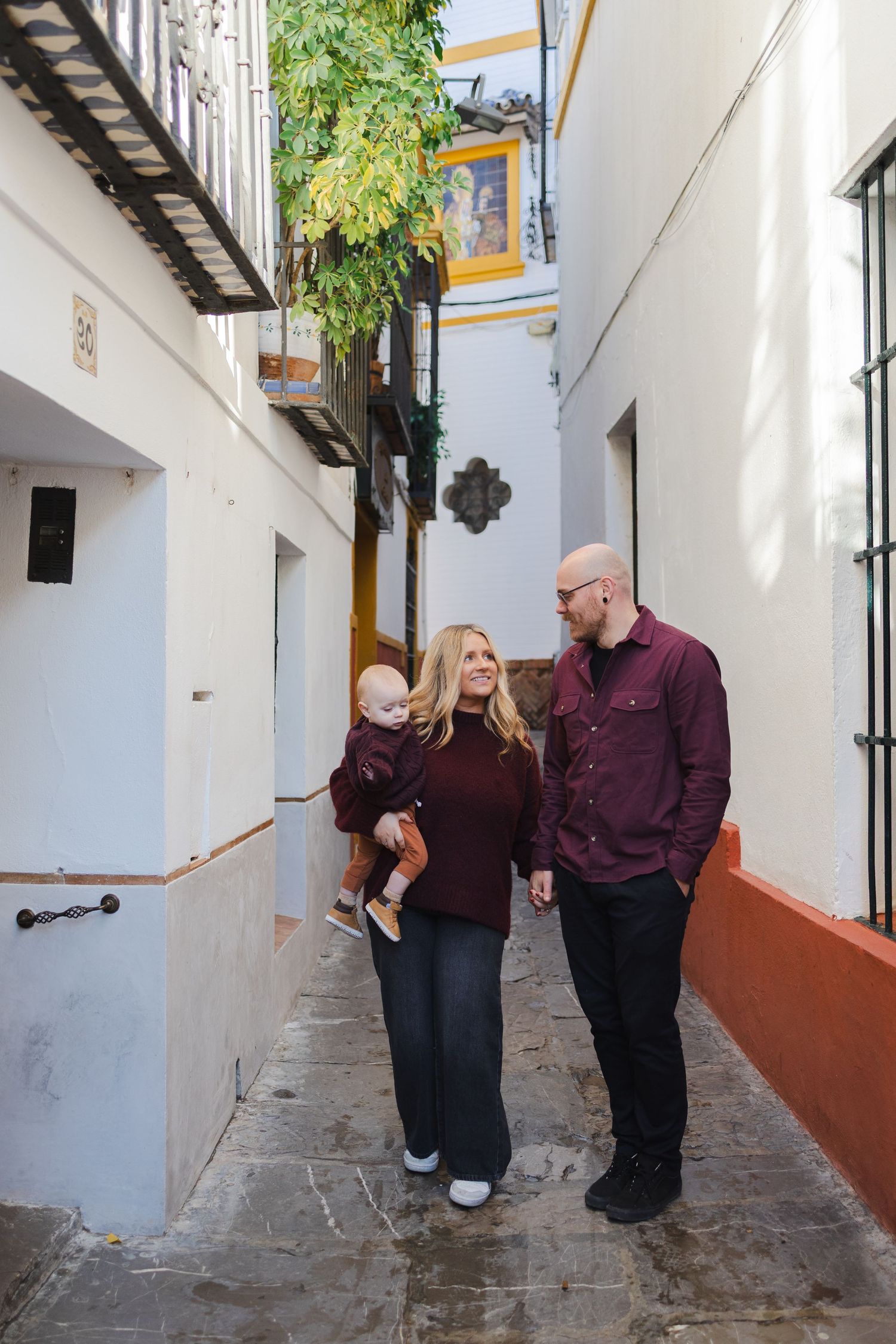 A family of three strolls through a charming narrow alley in Seville, Spain, dressed in coordinating burgundy outfits.