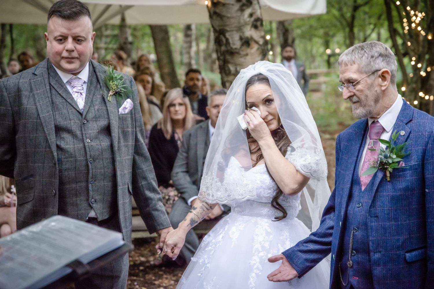 Emotional bride wiping tears during outdoor woodland wedding ceremony flanked by two men in suits.