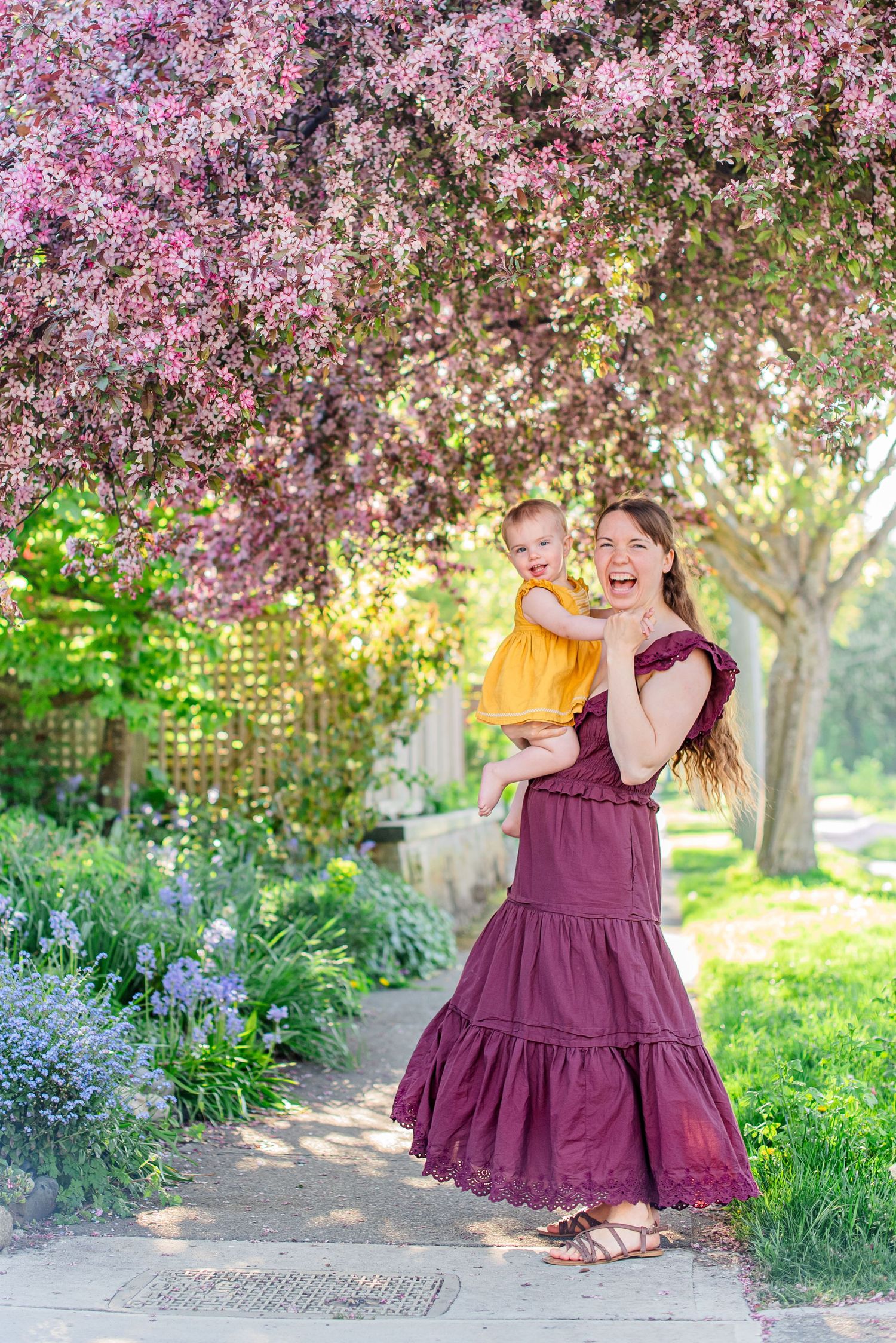 A joyful woman in a burgundy dress holds a baby in yellow under blooming pink trees in a lush garden.