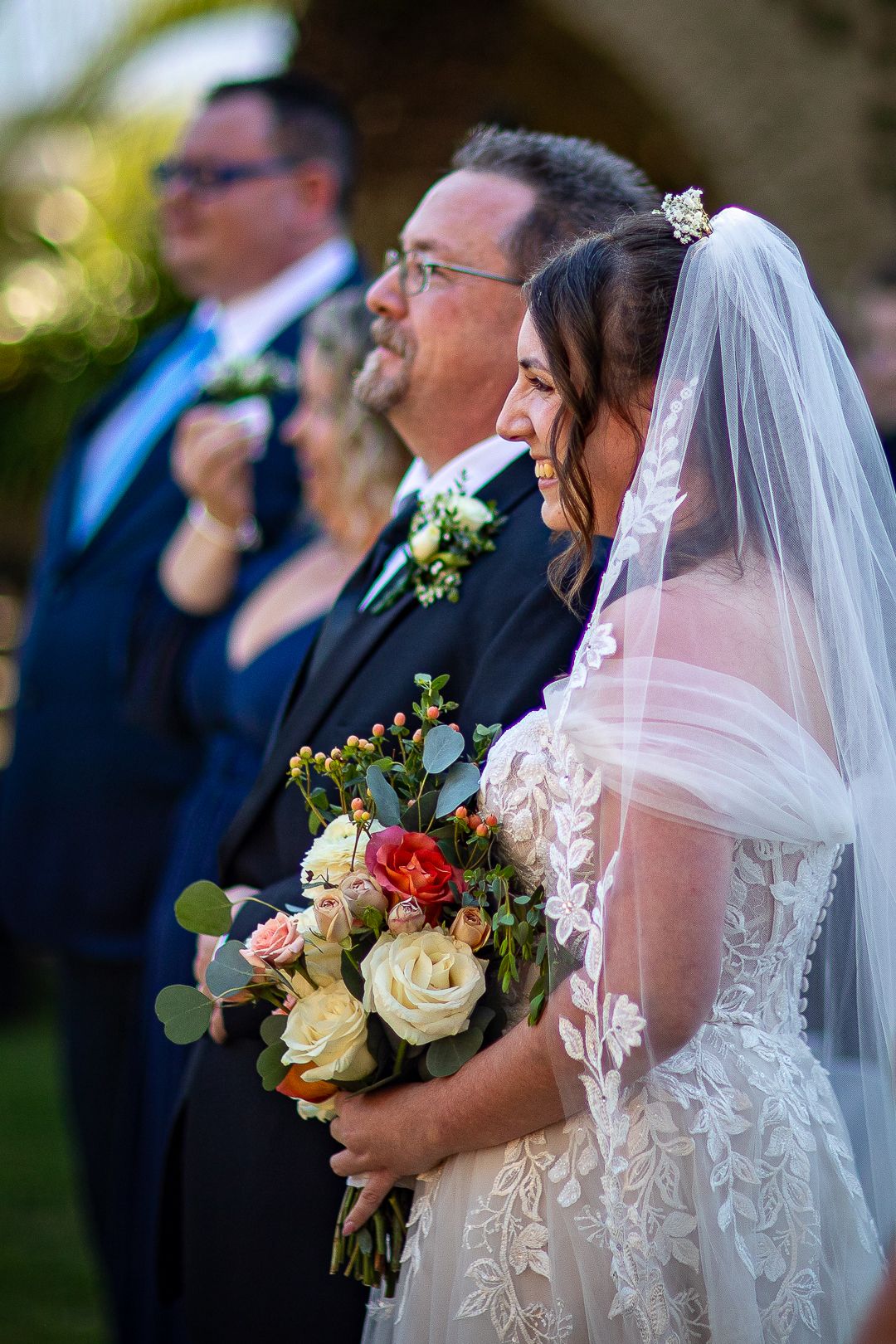 Bride in lace gown and veil holds colorful bouquet of roses at outdoor wedding ceremony with guests.