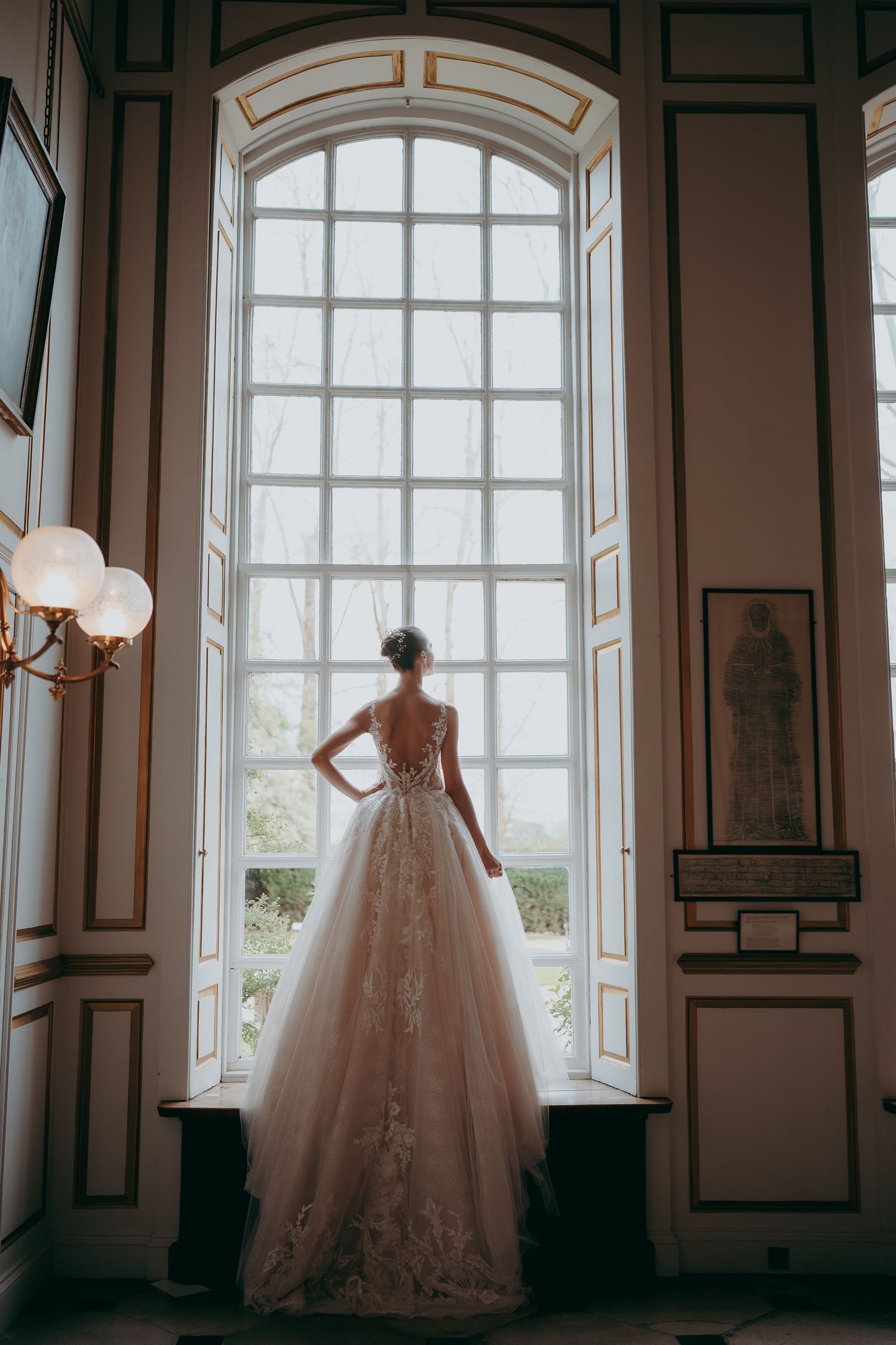 Bride in a backless lace ballgown stands before a grand arched window in an elegant, ornate room.