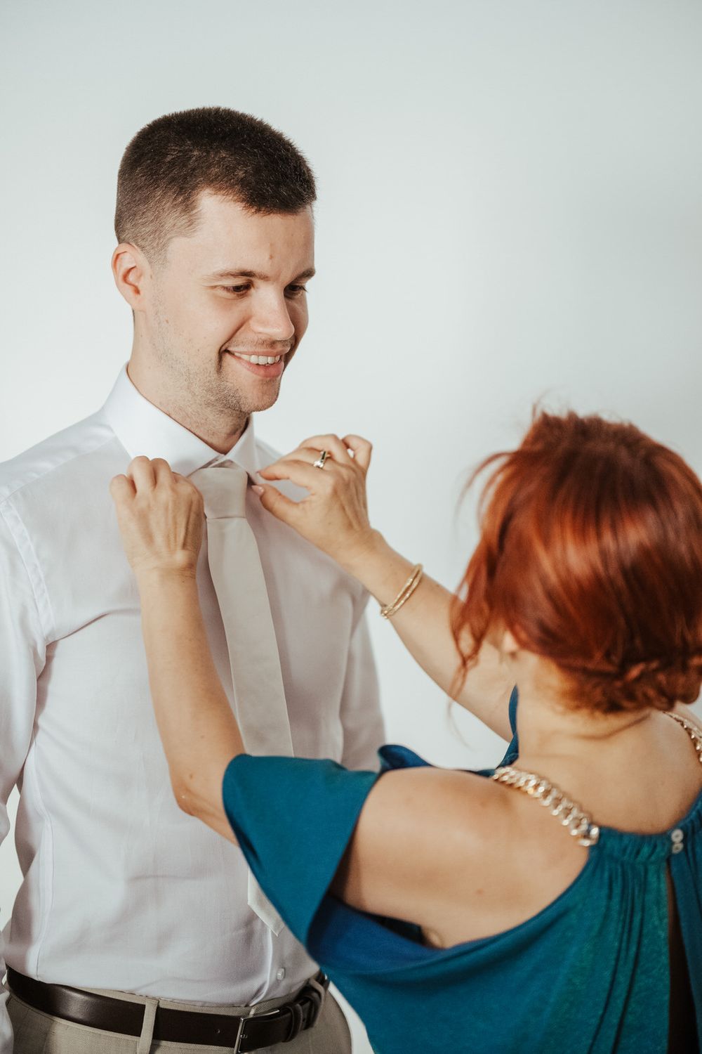 A smiling groom in a white shirt gets his tie adjusted by a redheaded woman in a teal dress against a light background.