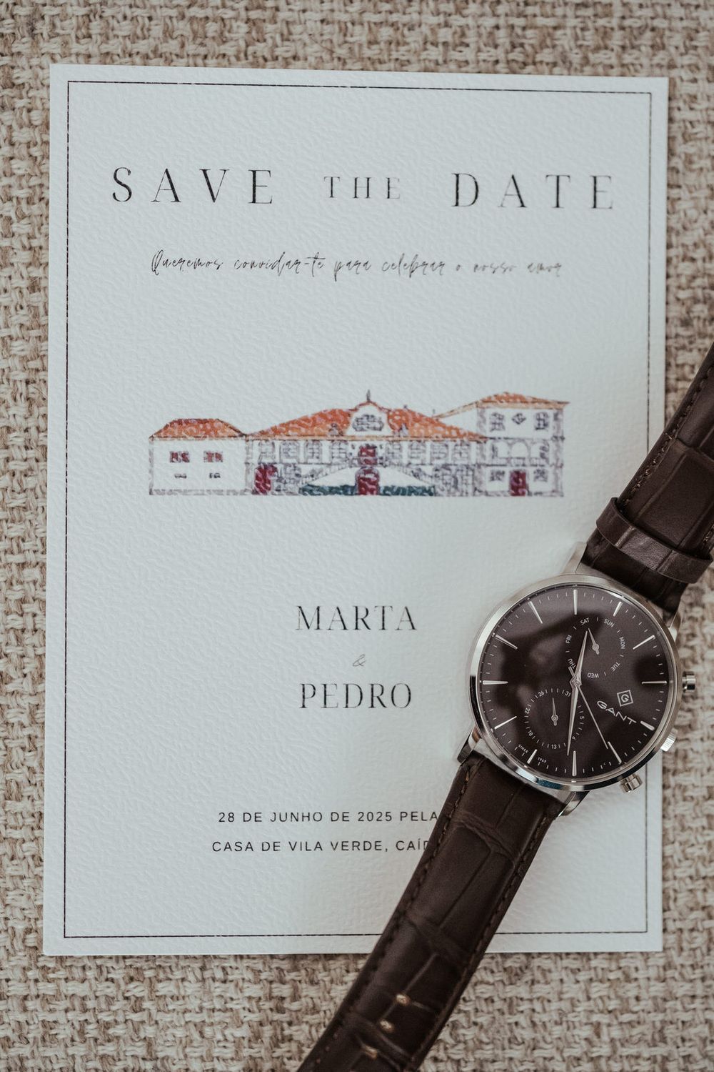 Elegant Save the Date card for Marta and Pedro on June 28, 2025, featuring a building illustration, with a brown leather watch on top.