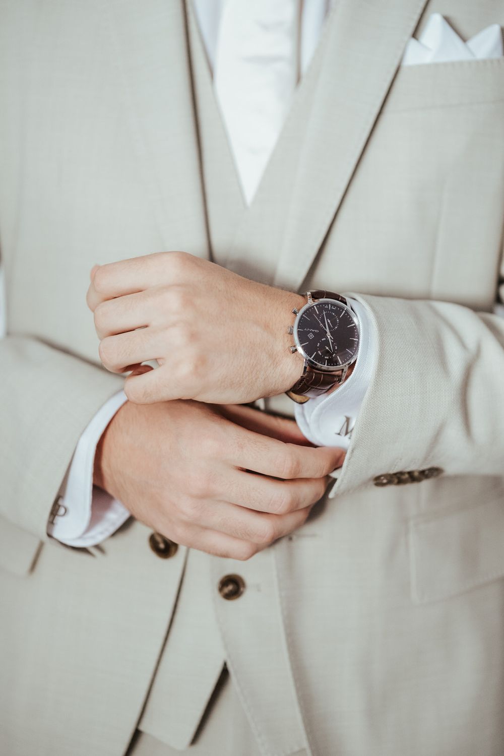 Man in beige suit adjusting cufflinks on white dress shirt while wearing an elegant brown chronograph watch.