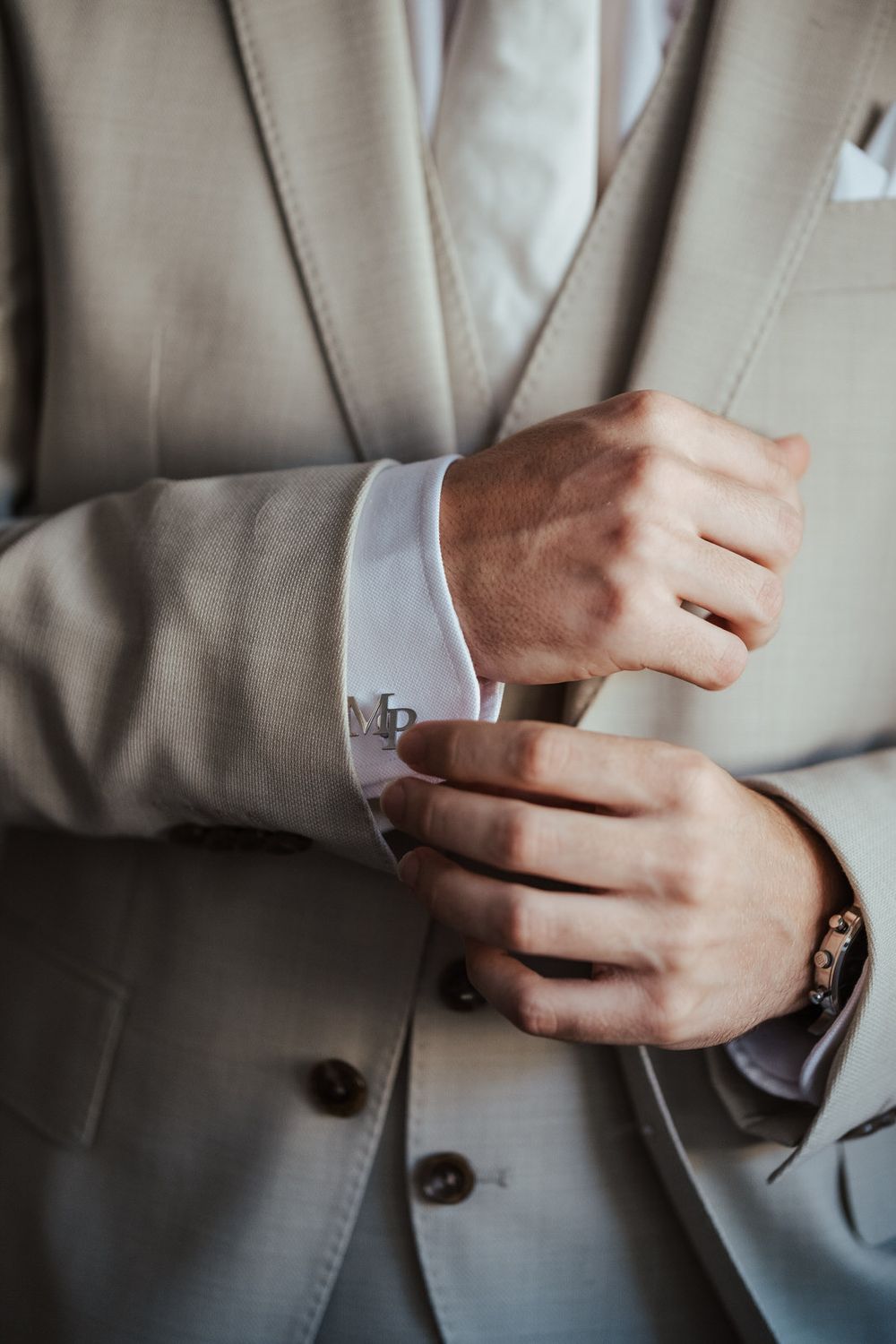 Man in beige suit adjusting monogrammed cufflinks on white dress shirt, wearing a bracelet and wedding ring.