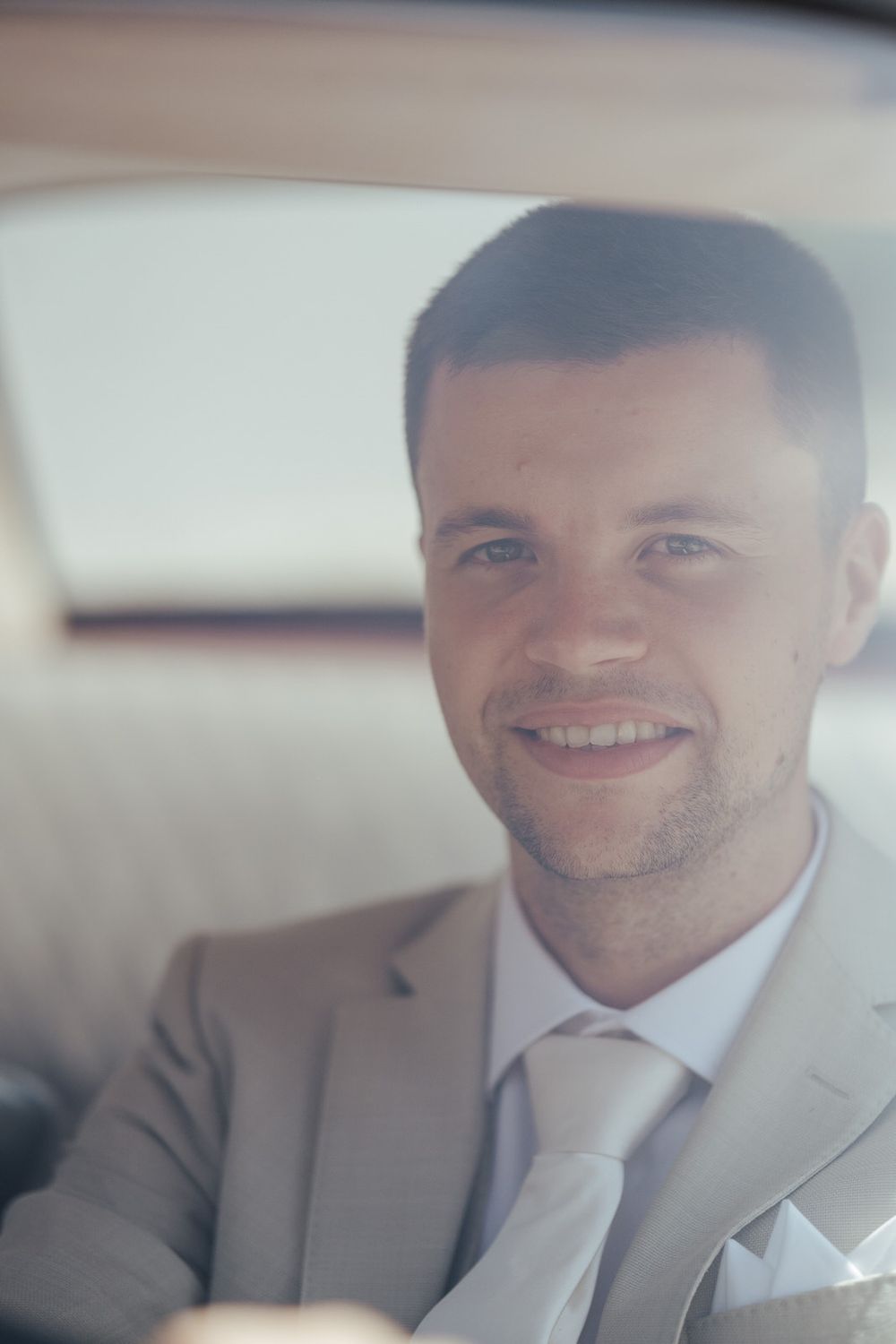 Smiling businessman in gray suit and tie sitting in car backseat, looking at camera through window.