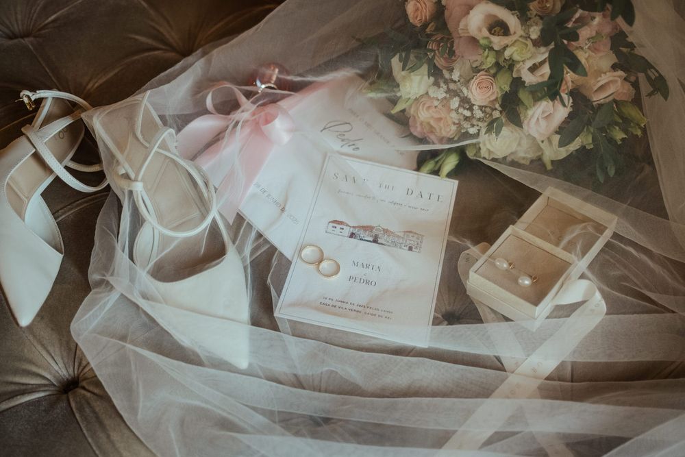 Elegant bridal flat lay featuring wedding invitation, pearl earrings, white heels, and a soft pink bouquet on tulle fabric.