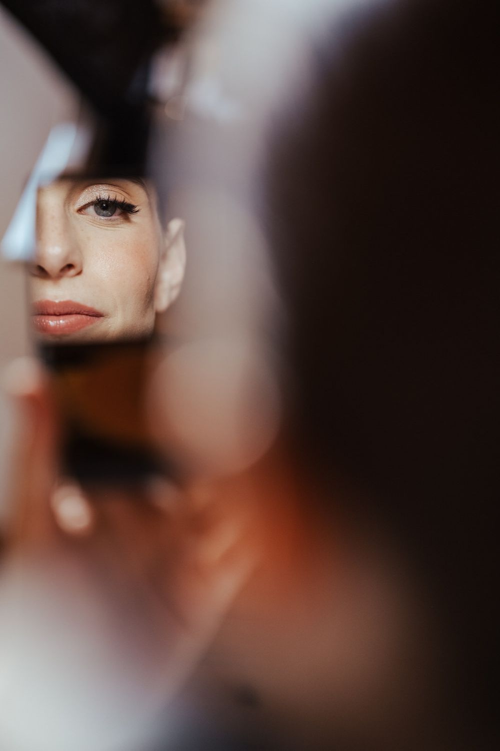 Woman's reflection in a small mirror showing natural makeup and dark hair in soft moody lighting.