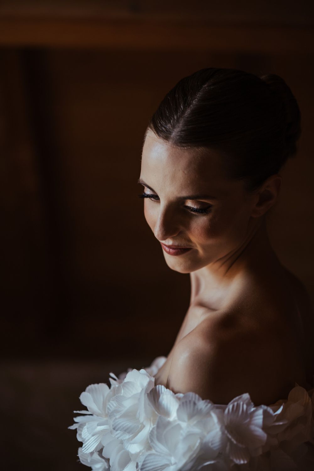 Elegant bride in white floral dress gazes down softly in dramatic moody lighting, hair in sleek updo.
