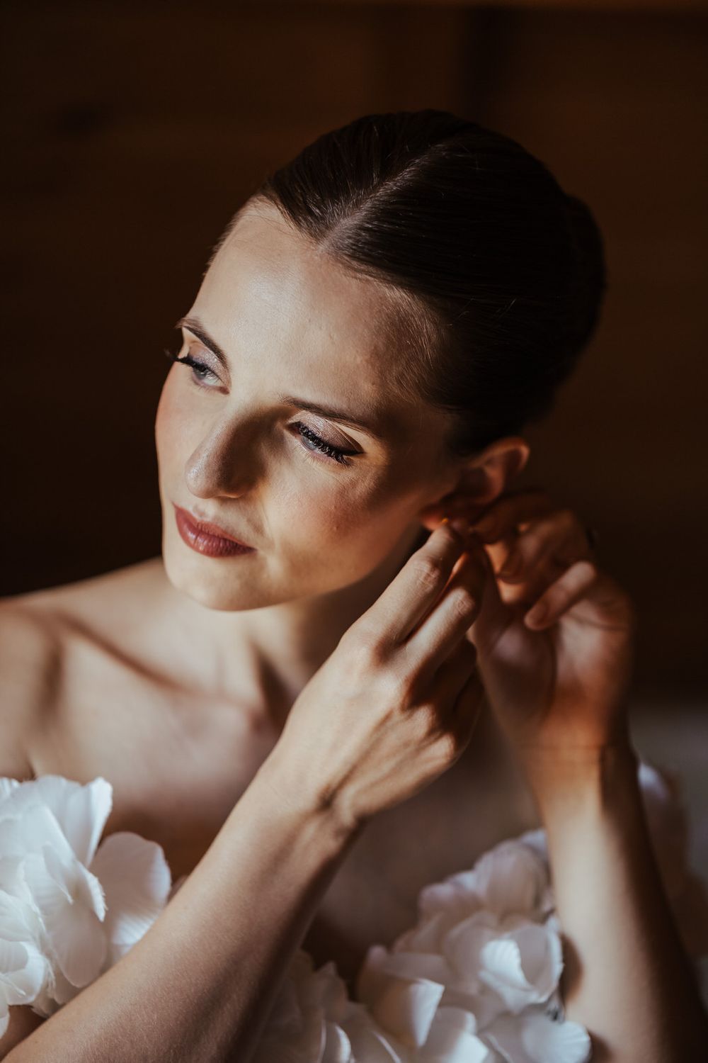 Elegant bride in a white floral dress adjusting her earring, with a sleek updo and natural makeup.