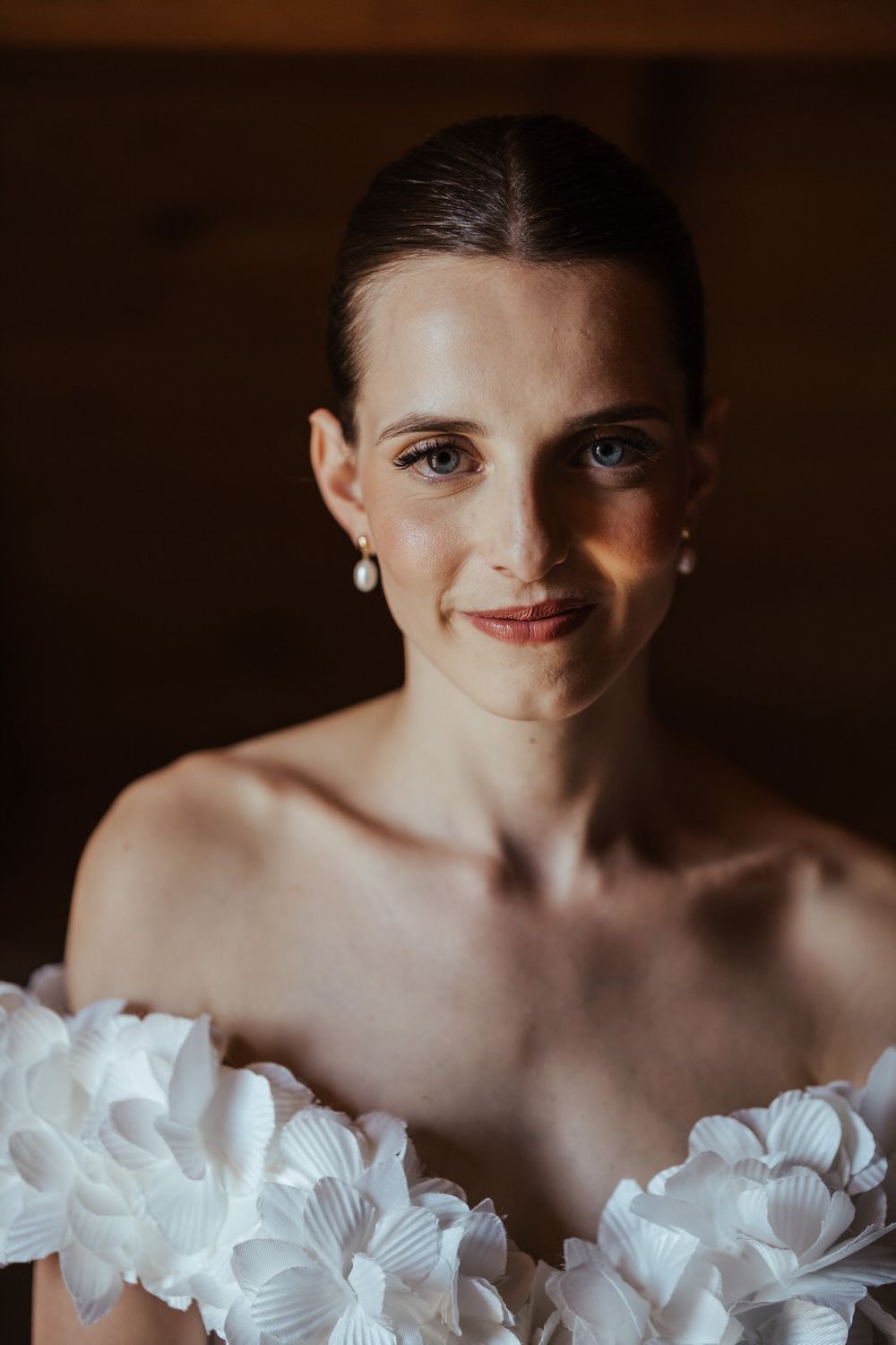 Elegant bride in off-shoulder white floral gown with pearl earrings, hair slicked back, soft smile, dark background.