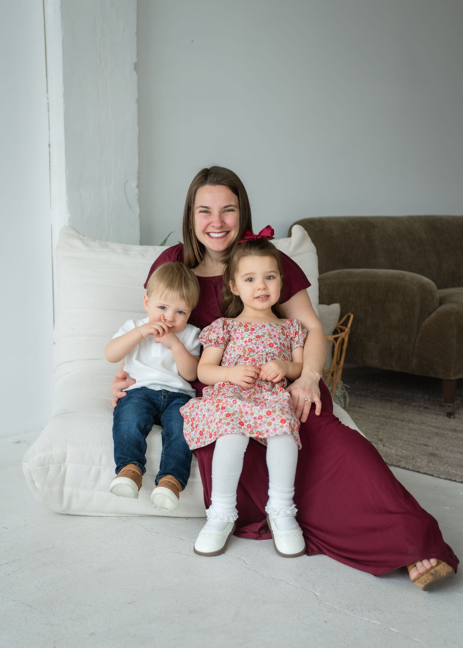 A smiling woman in a burgundy dress sits with two young children in a bright white studio setting.
