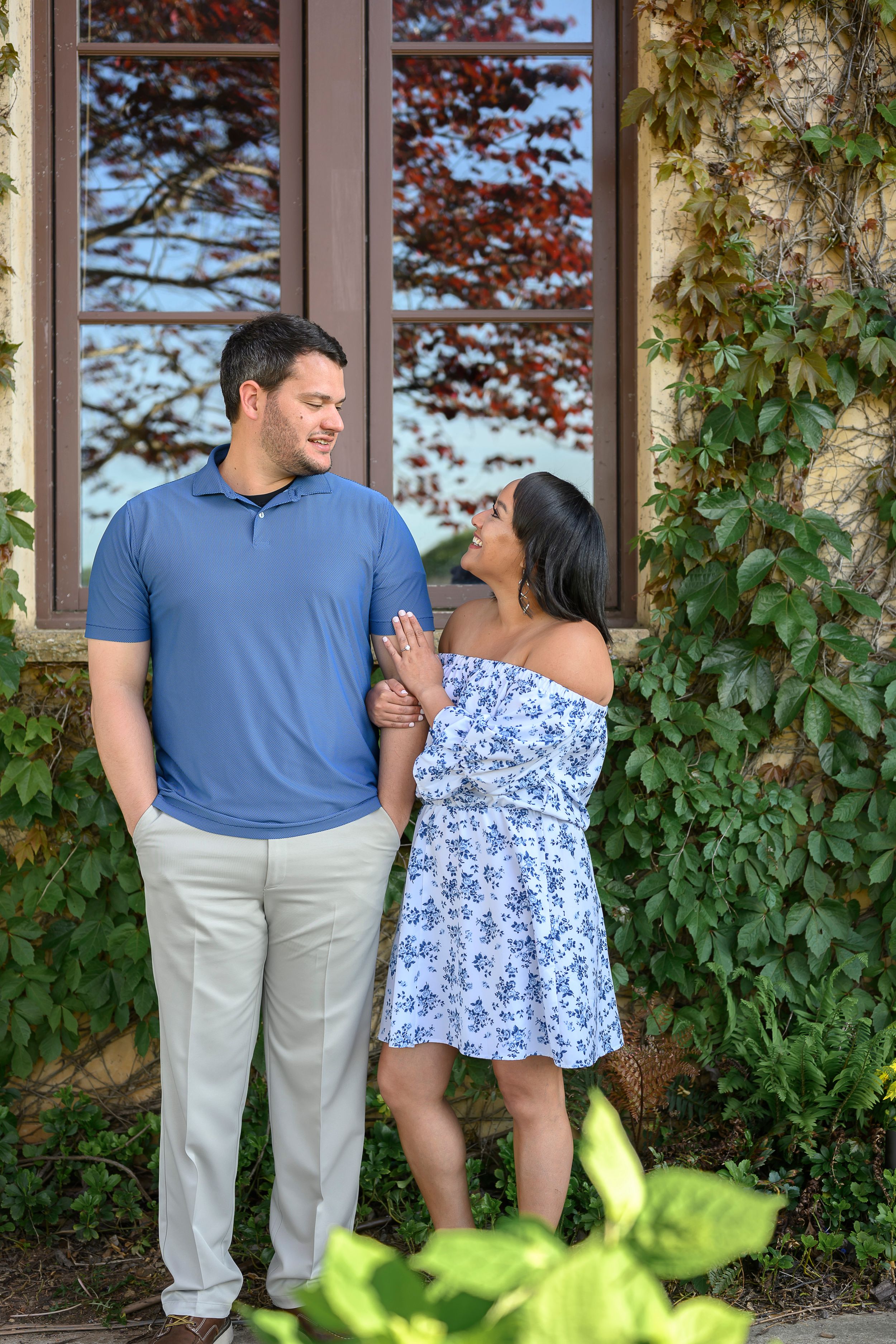 A couple smiles at each other outside a stone building covered in ivy, man in blue polo and woman in floral dress.