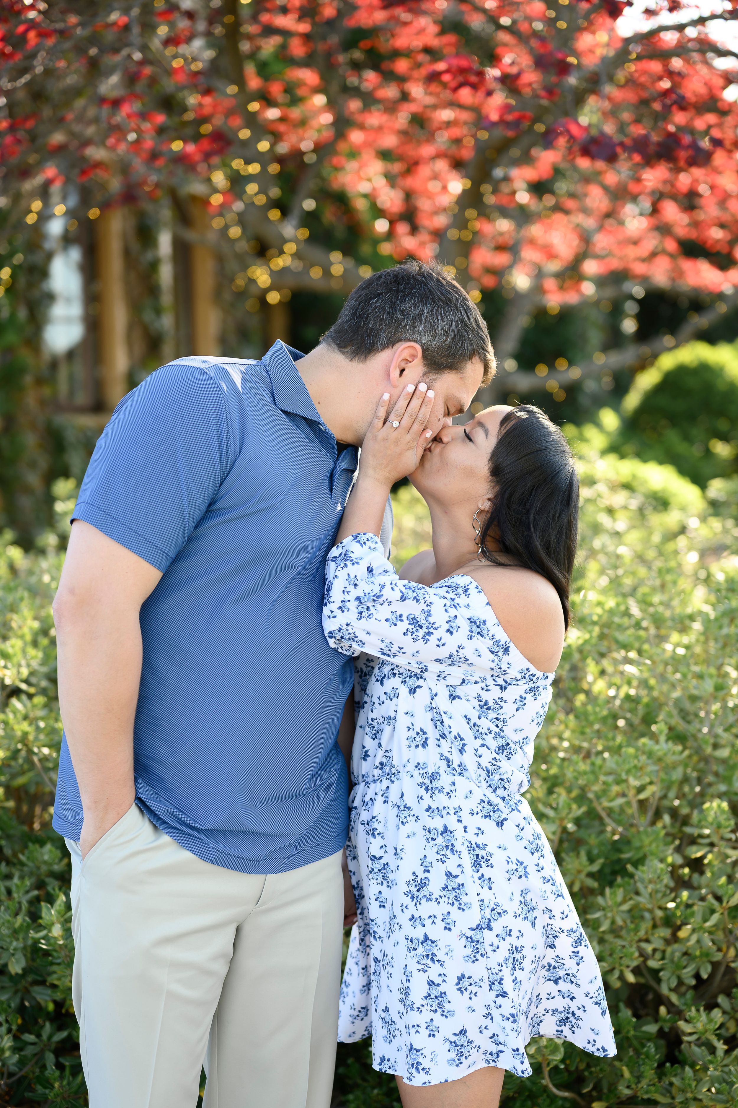 Couple sharing a romantic kiss outdoors, surrounded by lush green shrubs and vibrant red foliage in a garden setting.