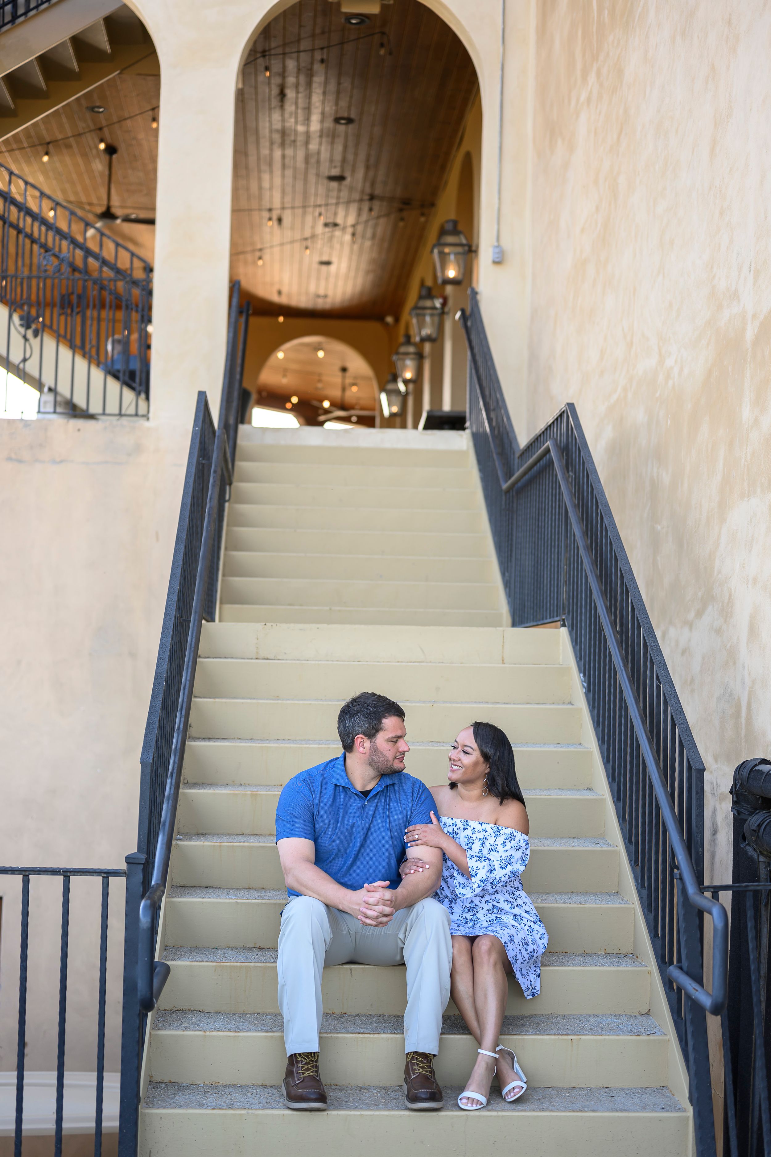 Couple sitting together on outdoor staircase, man in blue polo and woman in floral dress, smiling at each other.