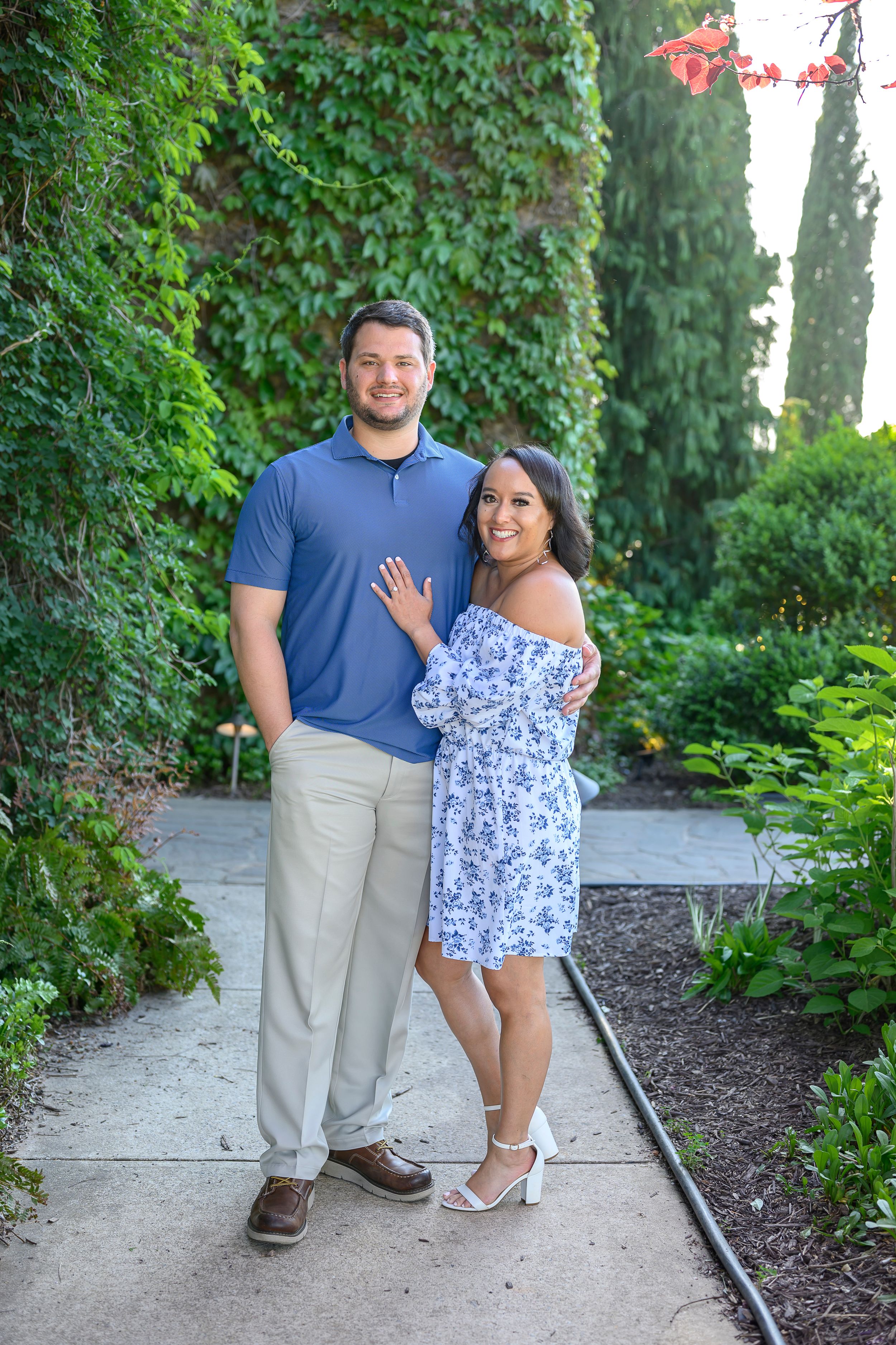 Couple posing on garden path, woman in floral dress showing engagement ring, man in blue polo, lush green ivy backdrop.