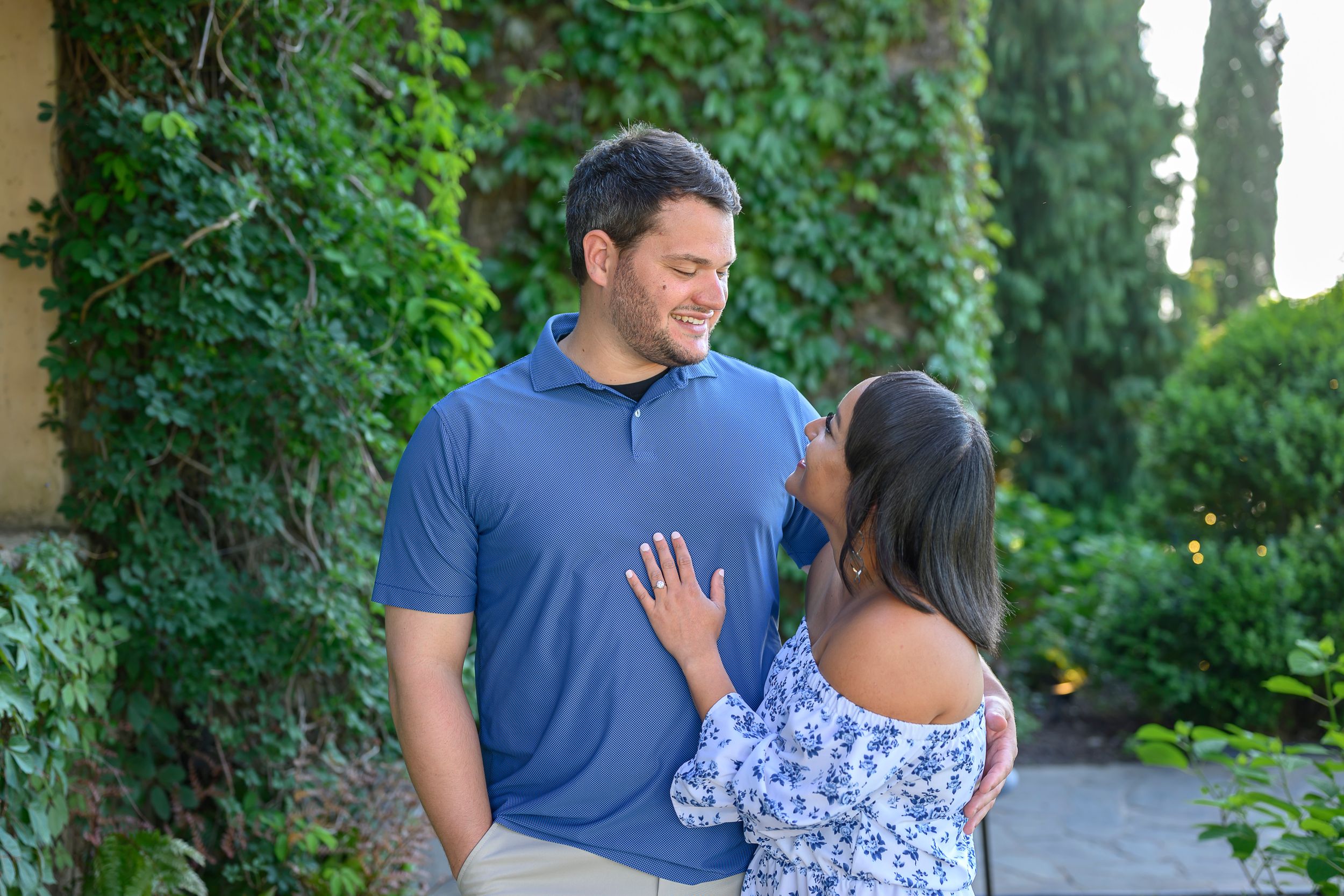 Couple sharing a romantic moment outdoors, man in blue polo smiling at woman in floral top, lush green ivy background.