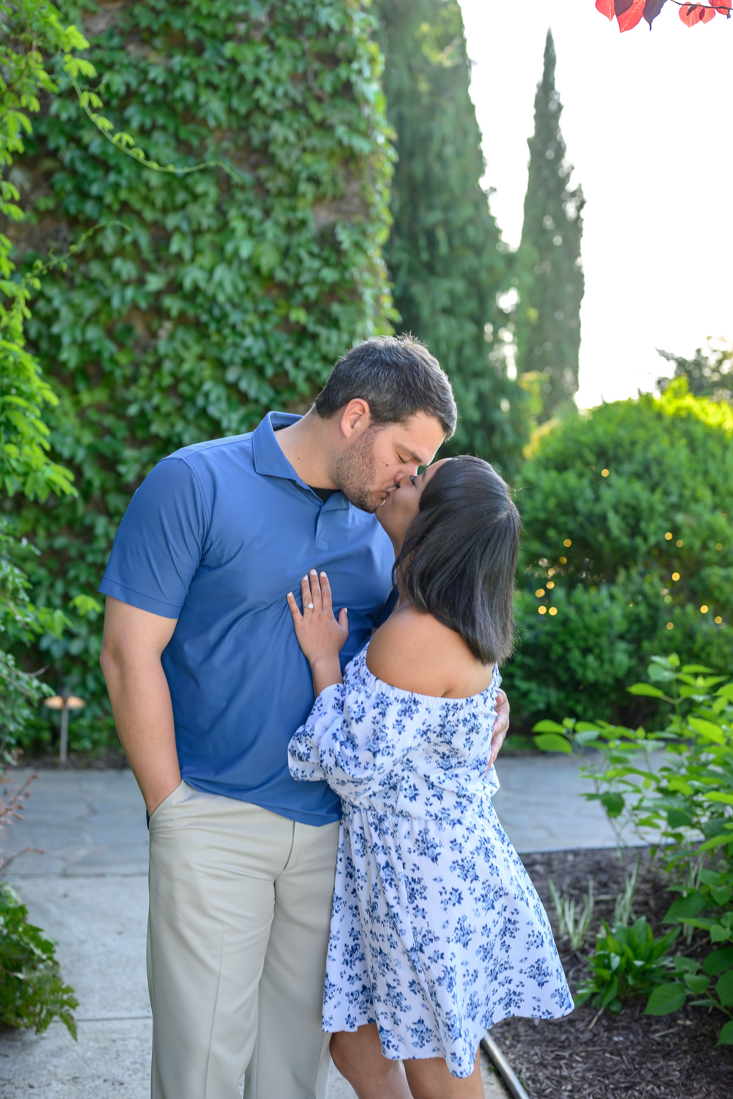 Couple sharing a romantic kiss in a lush garden, man in blue polo and woman in floral off-shoulder dress.