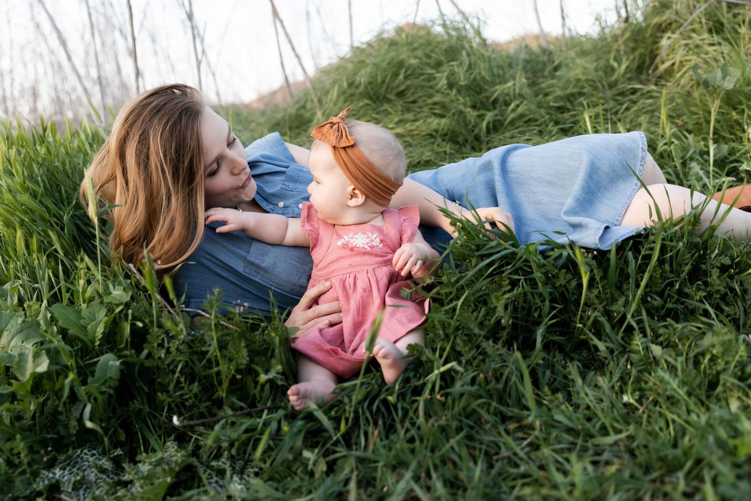 A loving mother lying in green grass gazes at her baby daughter in a pink dress during outdoor photos.