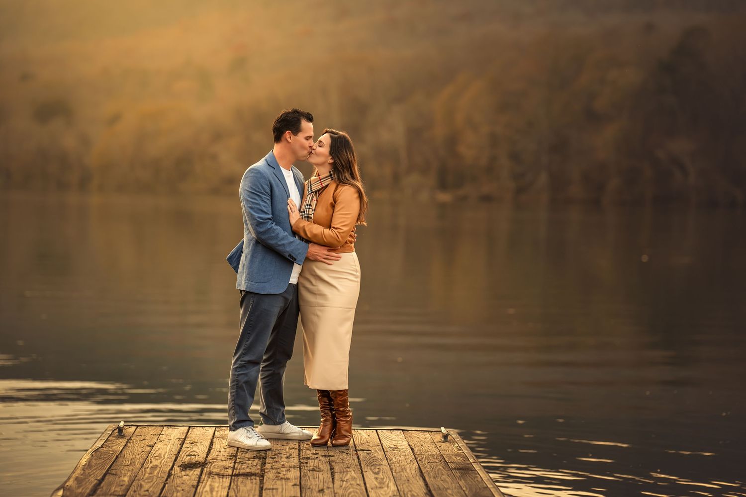 Couple kissing at sunset on a dock by a serene lake, woman in Burberry scarf and man in blue blazer.