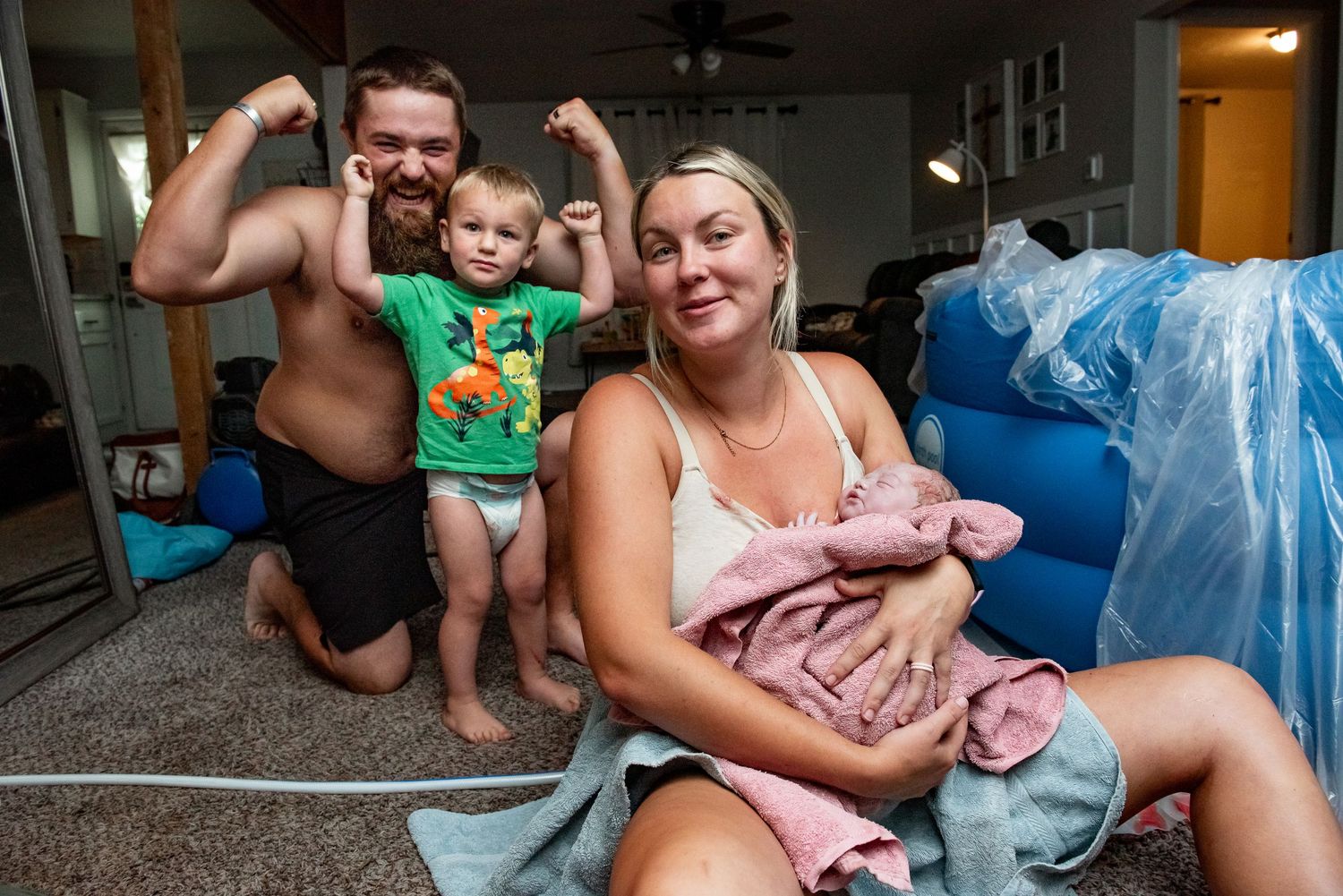 A family relaxes together on the floor of their living room with a baby wrapped in a pink blanket.