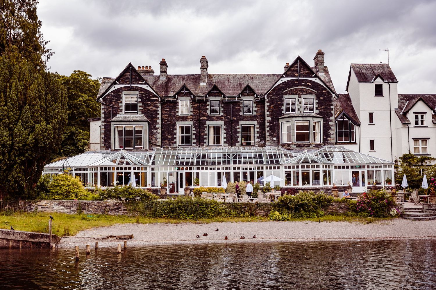 Victorian lakeside hotel with glass conservatory restaurant and wooden dock on calm grey waters surrounded by trees.