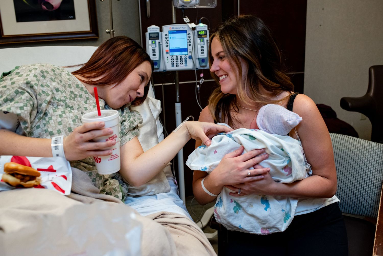 Two people sit together in a hospital room sharing joy while holding a swaddled newborn.