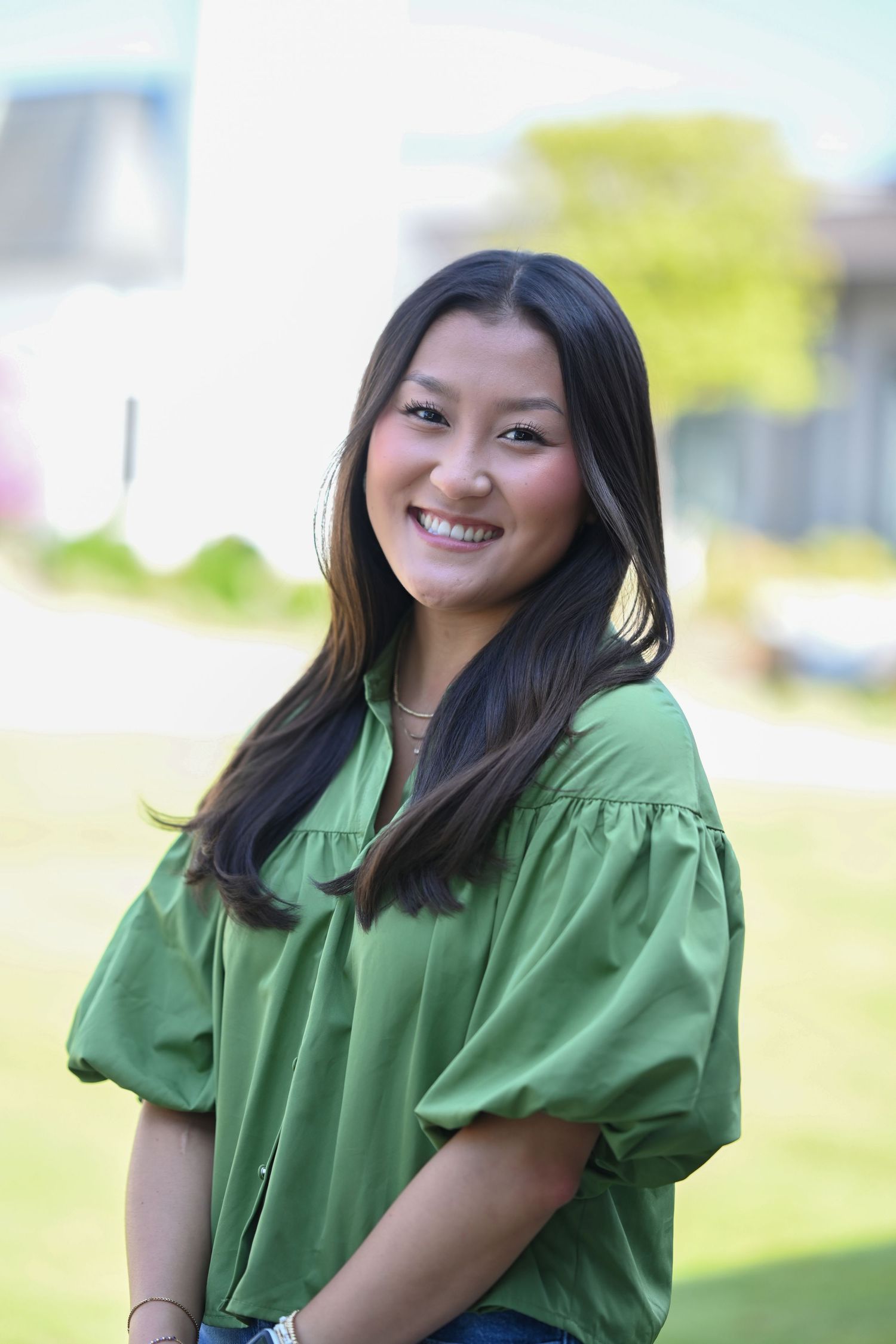 Smiling young woman with long dark hair wearing a green blouse, standing outdoors on a sunny day.