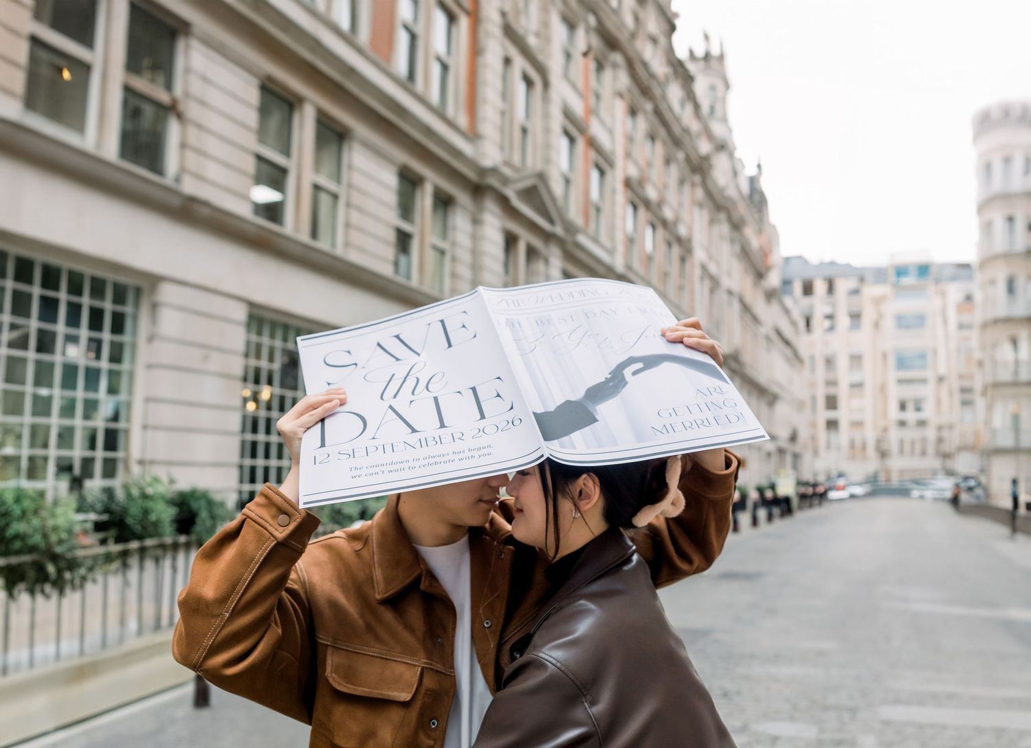 Couple kissing behind a Save the Date sign on a city street, wearing brown leather jackets.