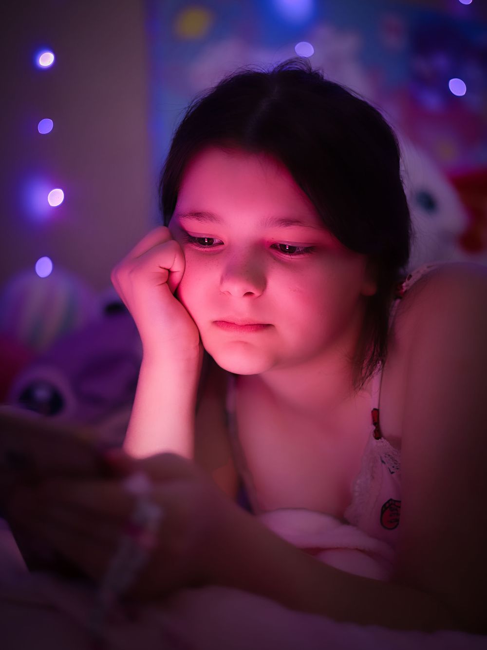 Young girl lying in bed illuminated by phone screen glow with colorful bokeh lights.