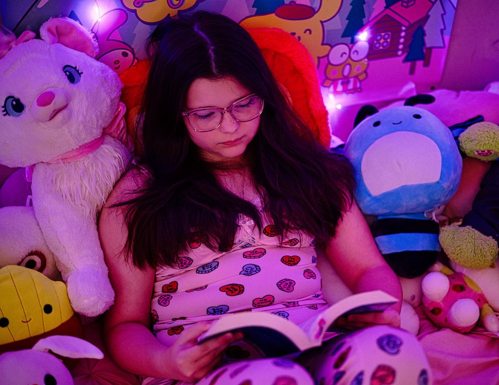 Girl in polka dot pajamas reading a book surrounded by colorful stuffed animals.