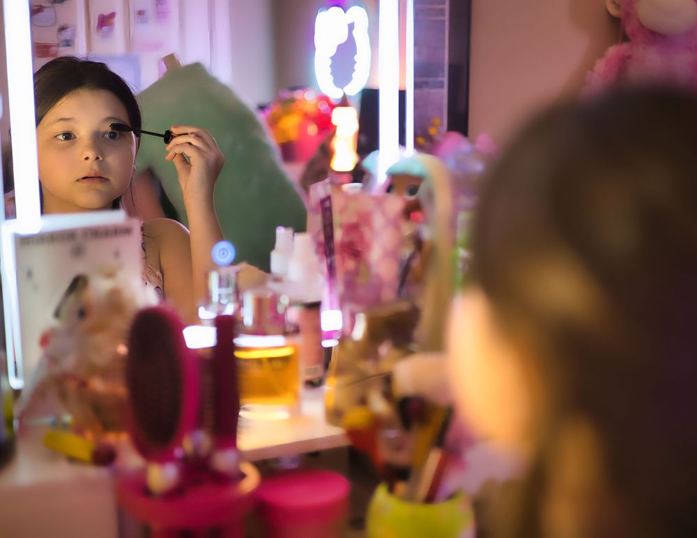 Young girl applying mascara at a vanity mirror surrounded by colorful beauty products.