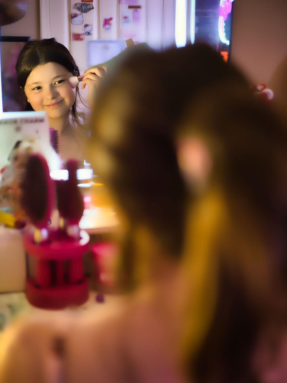Young girl smiling at her reflection in a vanity mirror with warm lighting.