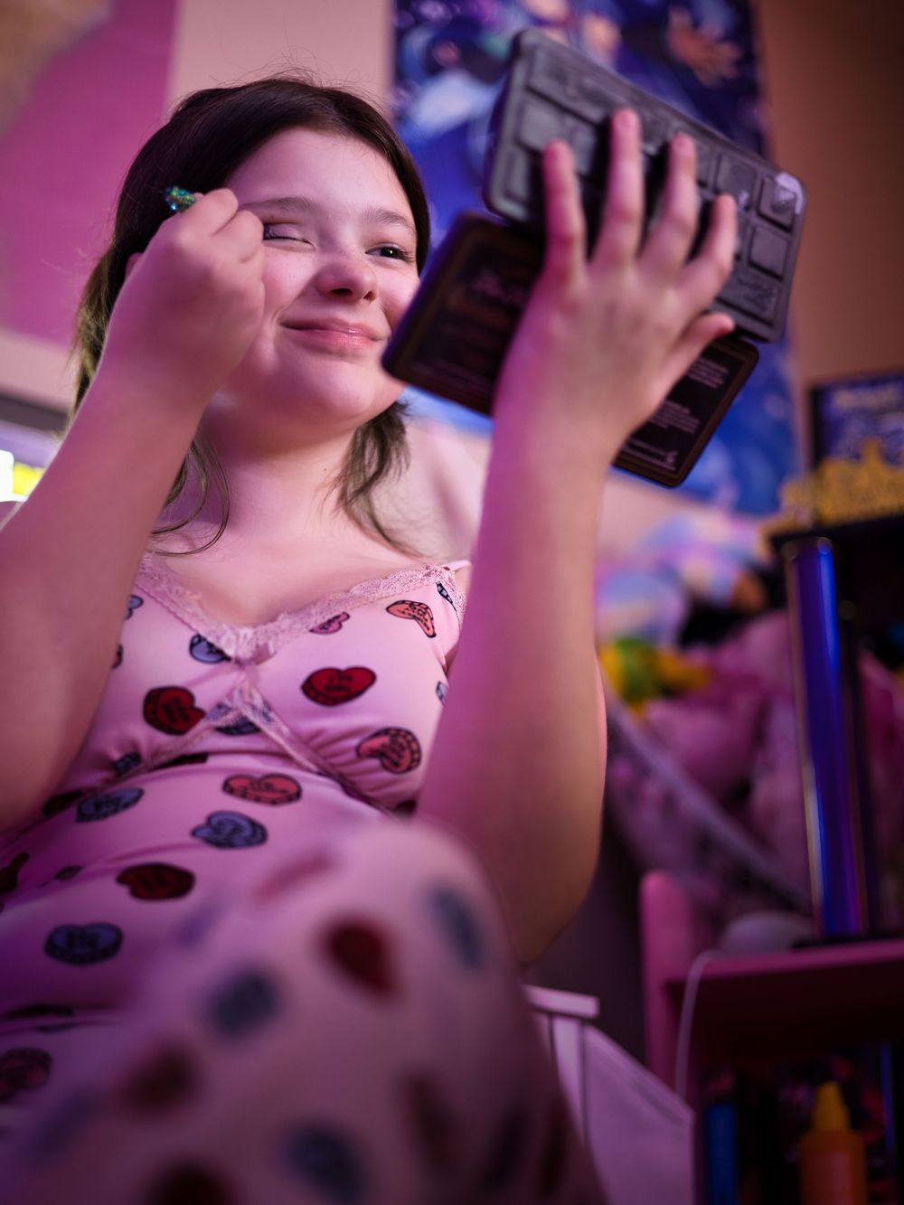 A young girl applies makeup while holding a compact mirror under pink neon lights.