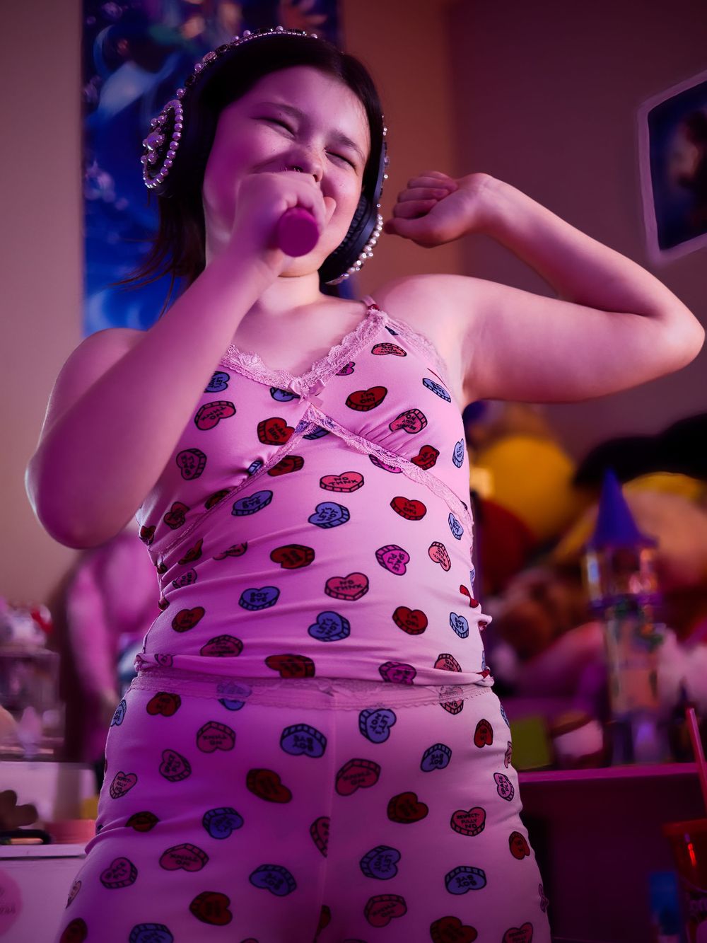 A young girl in a pink polka dot outfit poses playfully under pink neon lights.