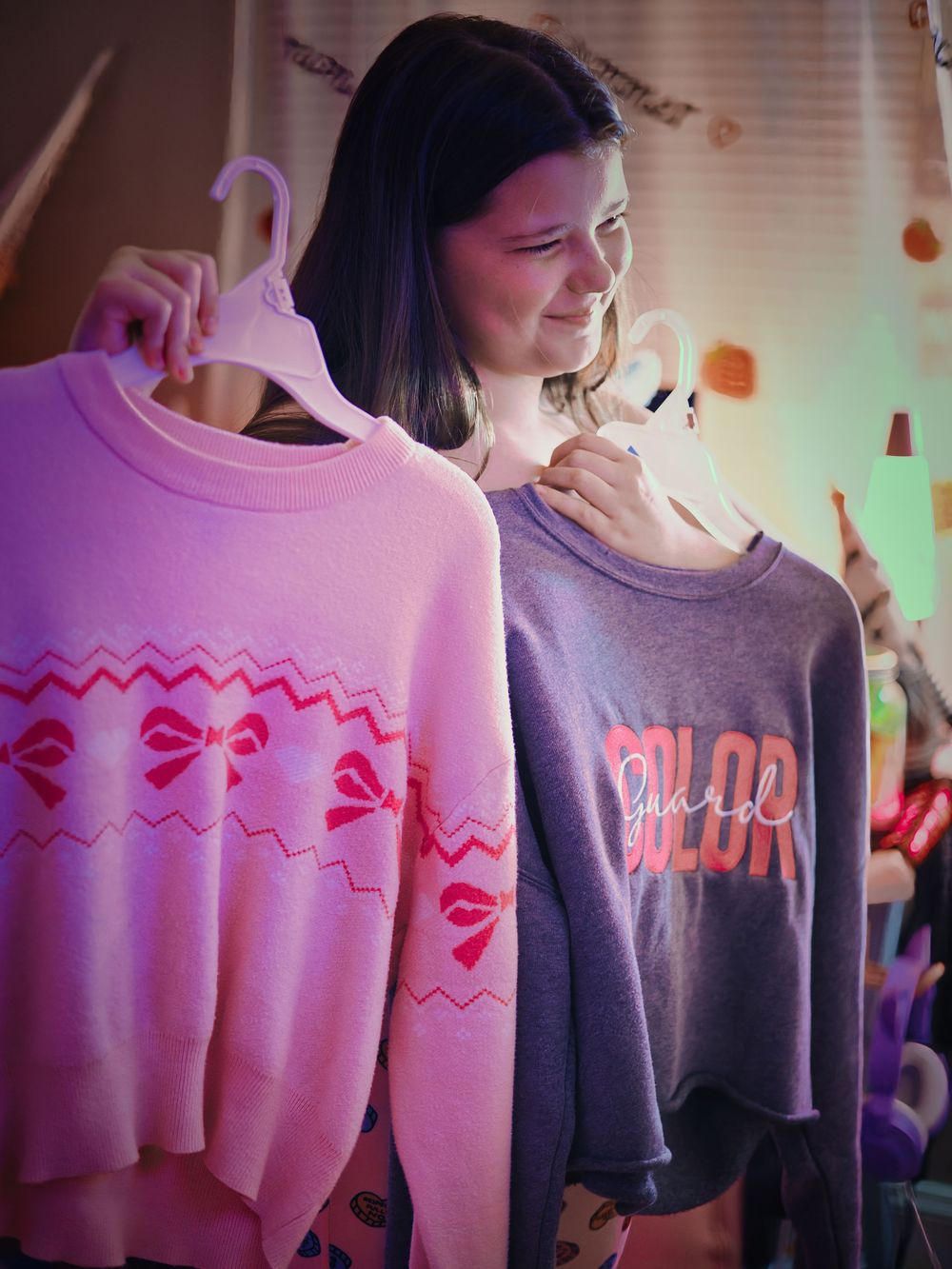 A young woman smiles while holding two colorful sweaters on hangers in a pink-lit room.