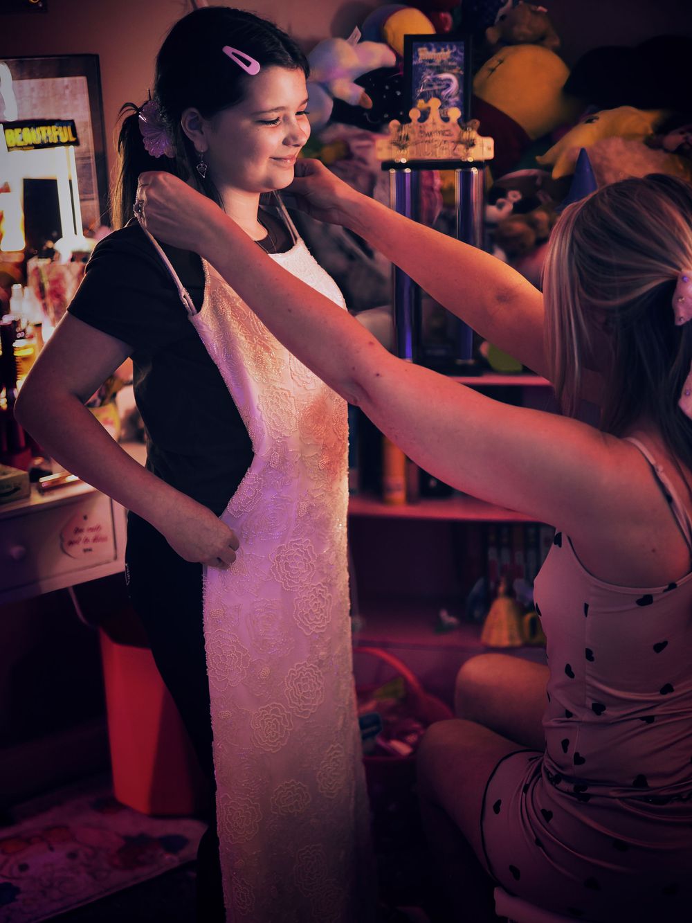 A woman fastens a necklace on a young girl holding a white dress in a warmly lit room.