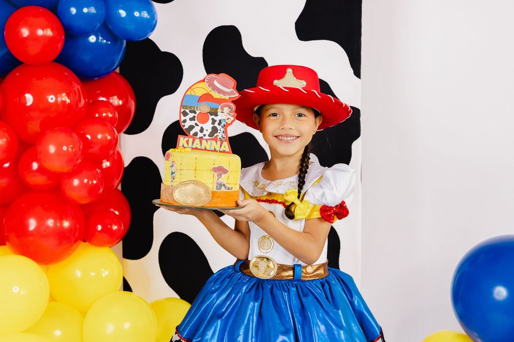 Girl in Jessie Toy Story costume holding birthday cake at colorful cowprint party with red, blue, yellow balloons.