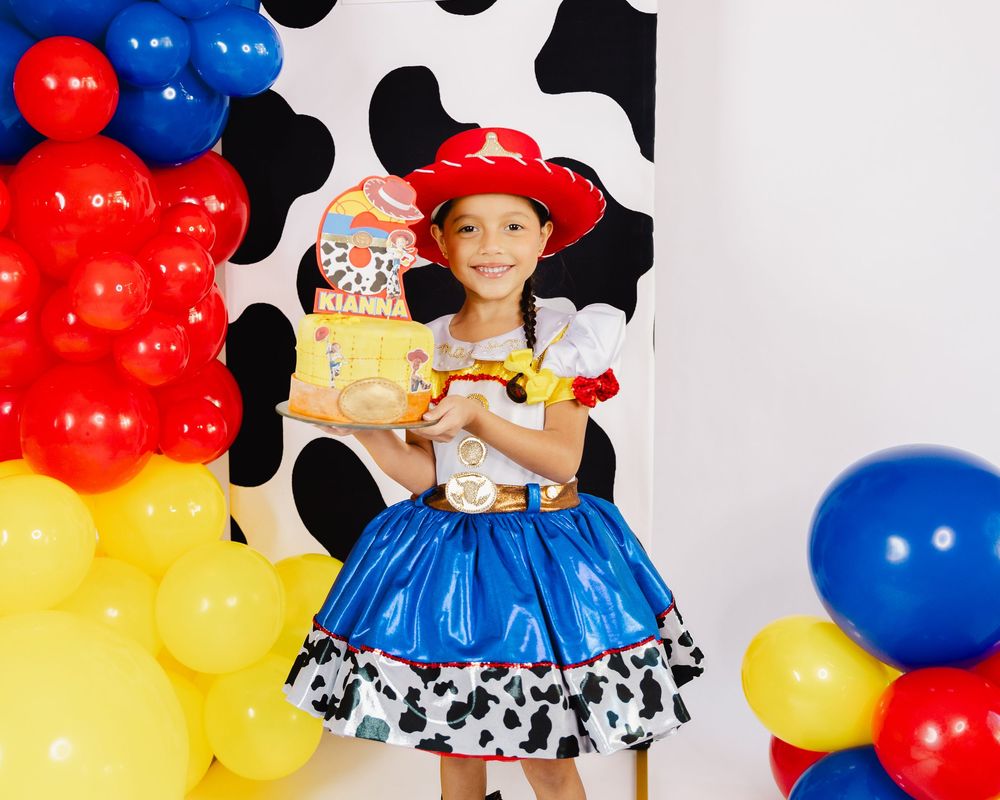 Girl in Jessie Toy Story costume holds gift box at colorful balloon party backdrop with cow print decor.