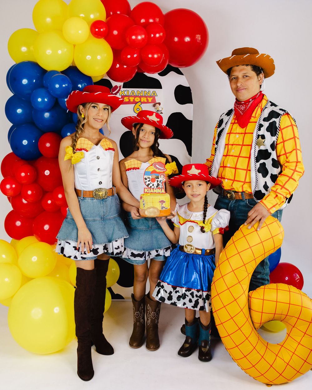 Family dressed in Toy Story Woody and Jessie costumes posing in front of colorful balloon arch at birthday party.