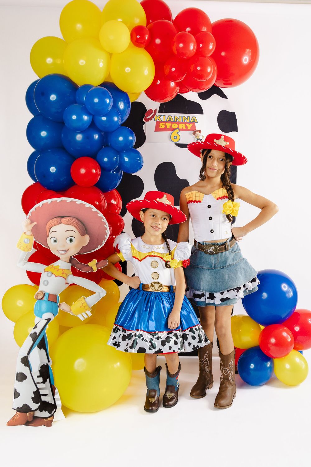 Two girls in Jessie Toy Story costumes pose with a cardboard cutout beside colorful balloons at a themed birthday party.