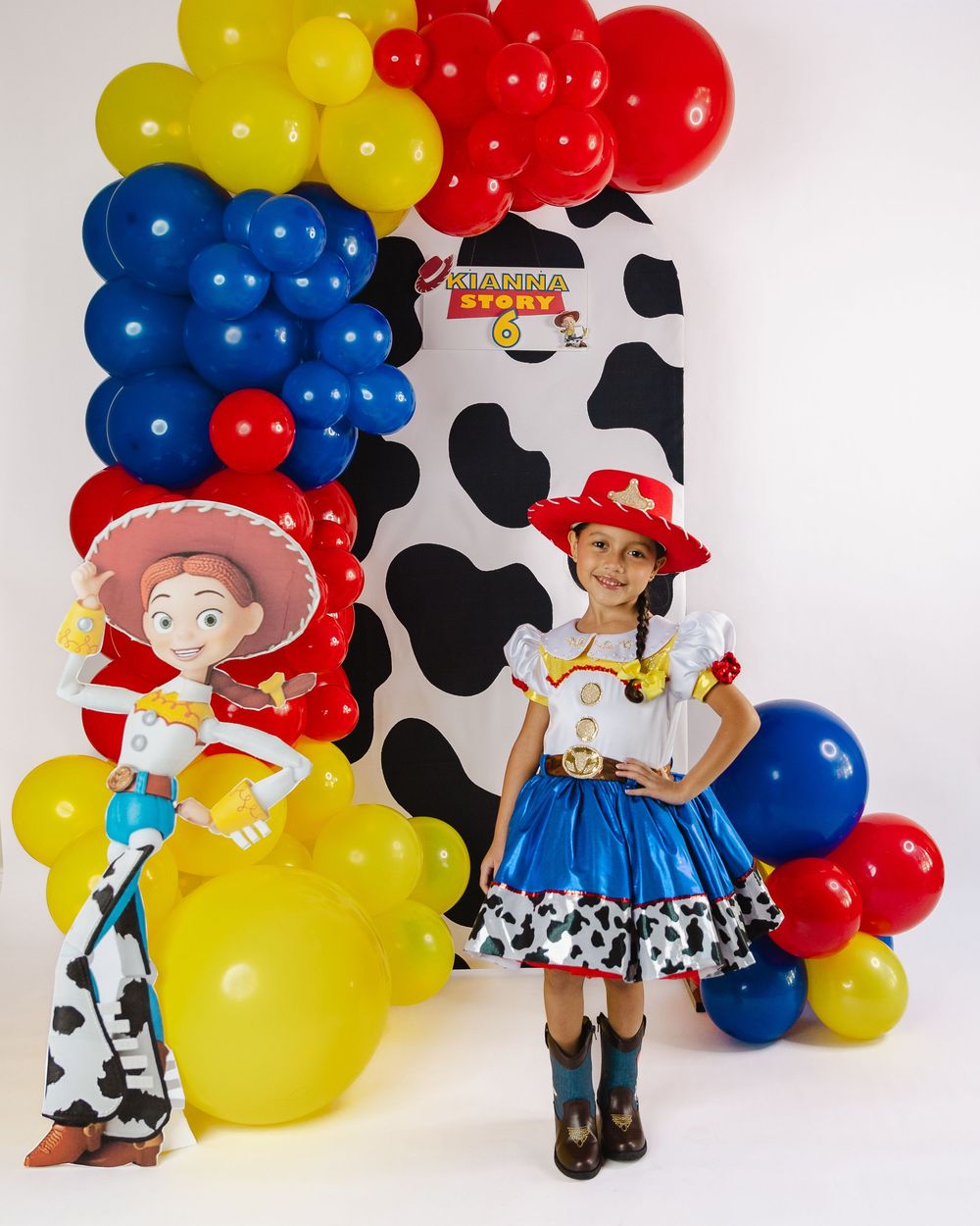 Girl in Jessie Toy Story costume poses beside character cutout with colorful balloon arch at themed birthday party.