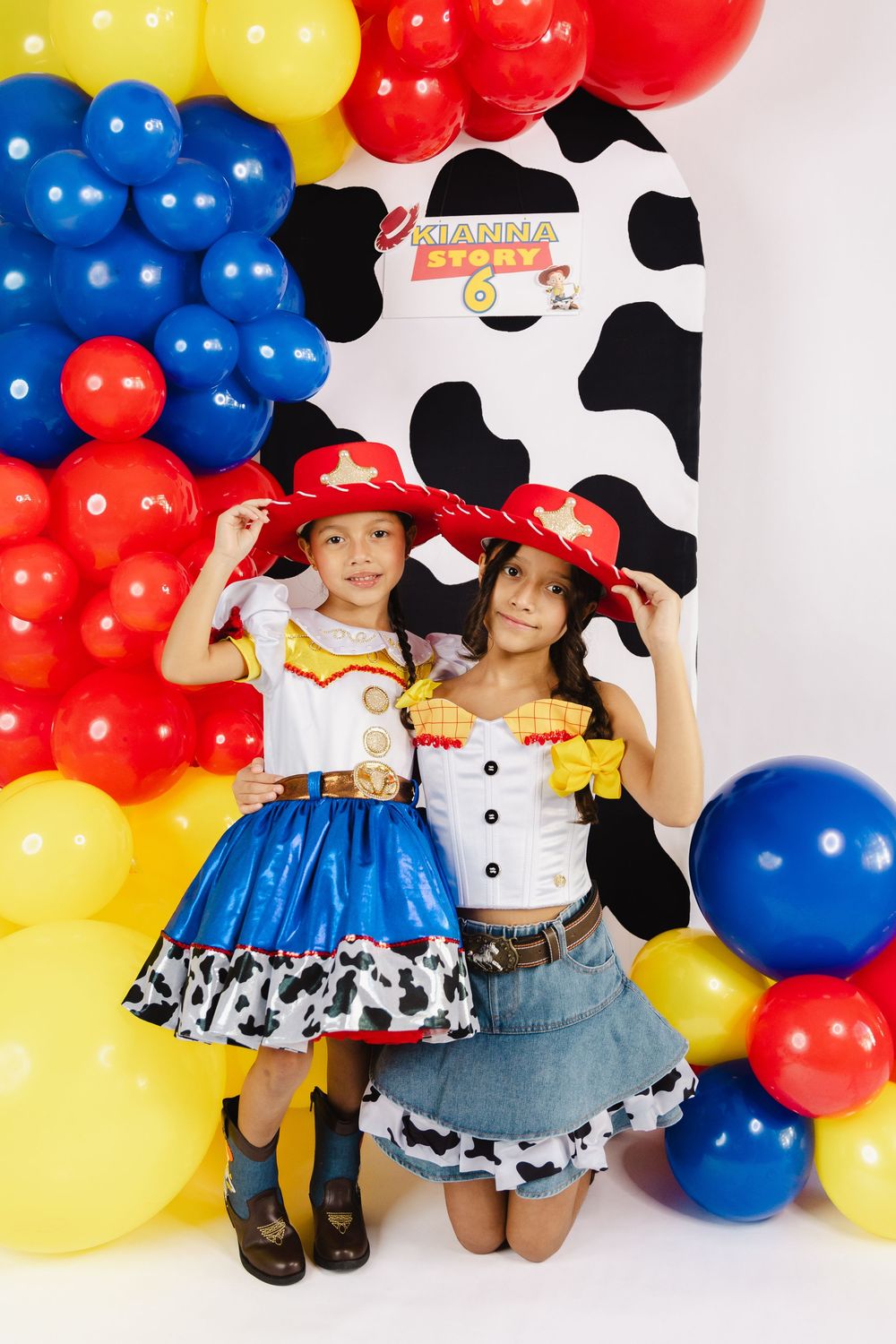 Two girls in Jessie Toy Story costumes pose at a Kianna Story 6th birthday party with colorful balloon arch backdrop.