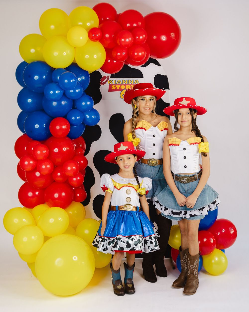 Three females in Toy Story Jessie costumes pose before a red, blue, and yellow balloon arch at a themed party.