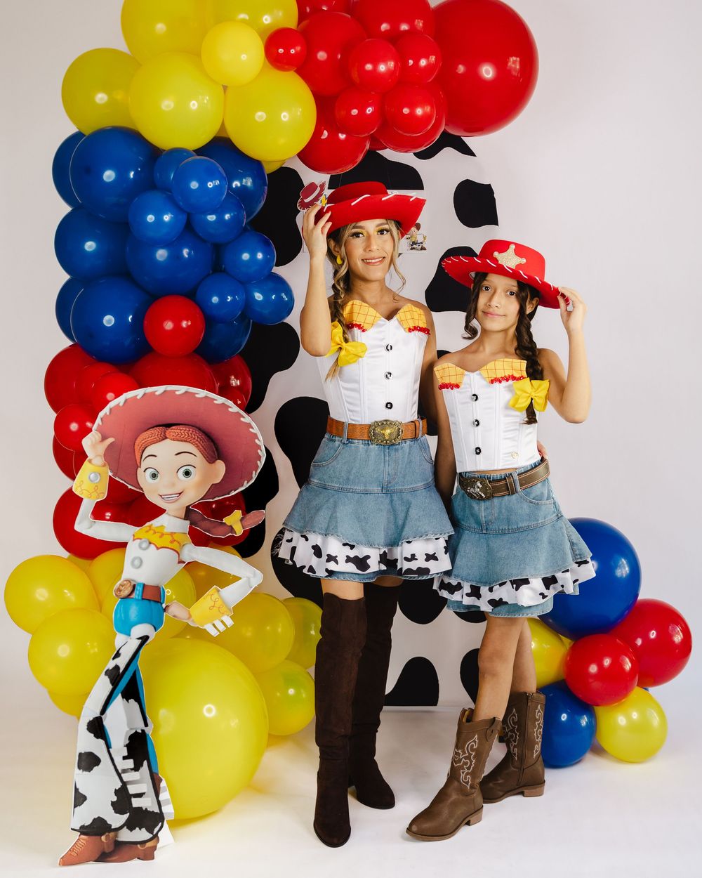 Two women dressed as Jessie from Toy Story pose with a cardboard cutout at a colorful balloon party backdrop.