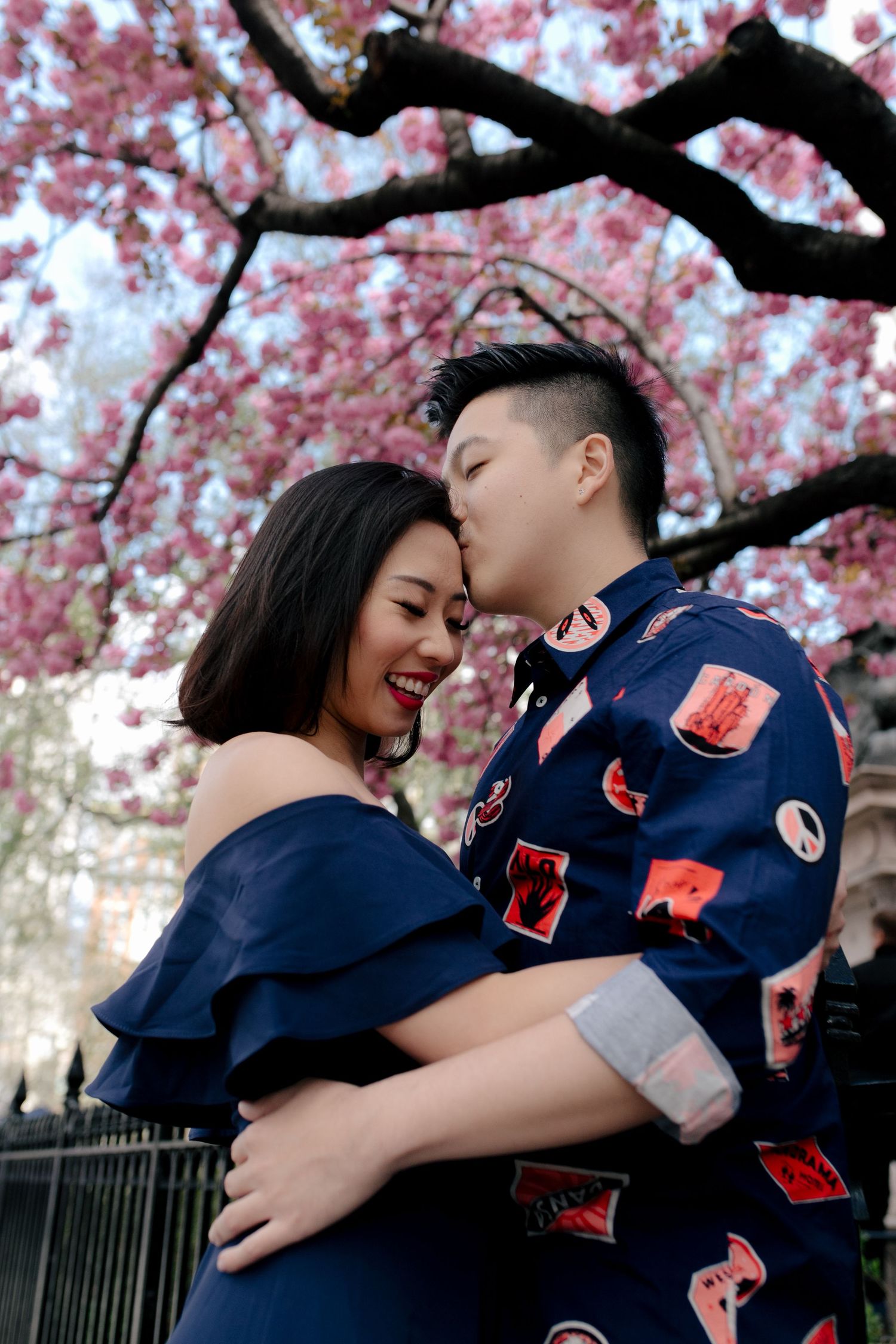 Couple embracing under pink cherry blossom tree, wearing matching navy blue outfits, sharing a romantic kiss outdoors.