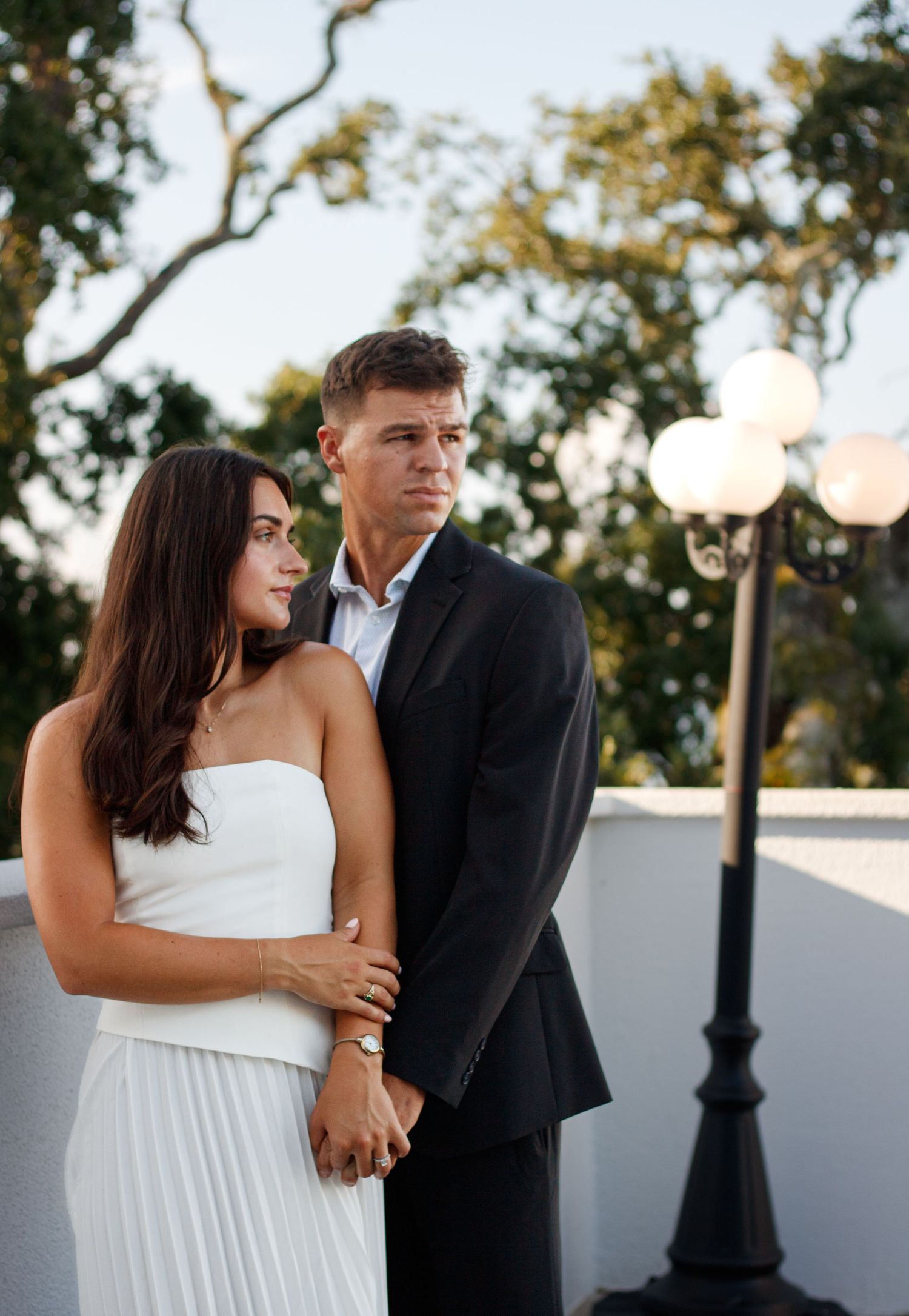 A woman in a white dress and a man in a dark suit stand close together beside a lamp post at sunset.